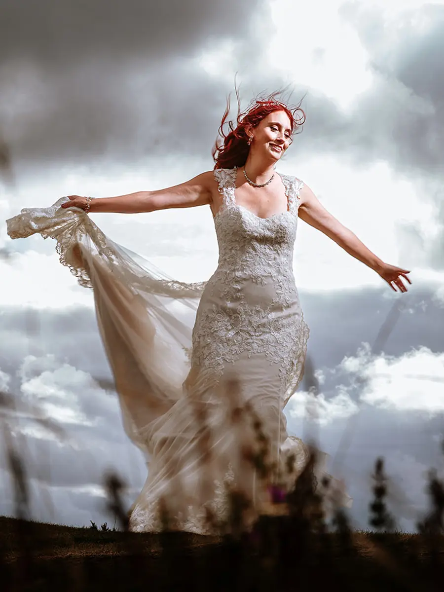 Bride with red hair arms outstretched holding her dress in the wind against a dramatic cloudy sky — documentary wedding photography by Gary Phillip
