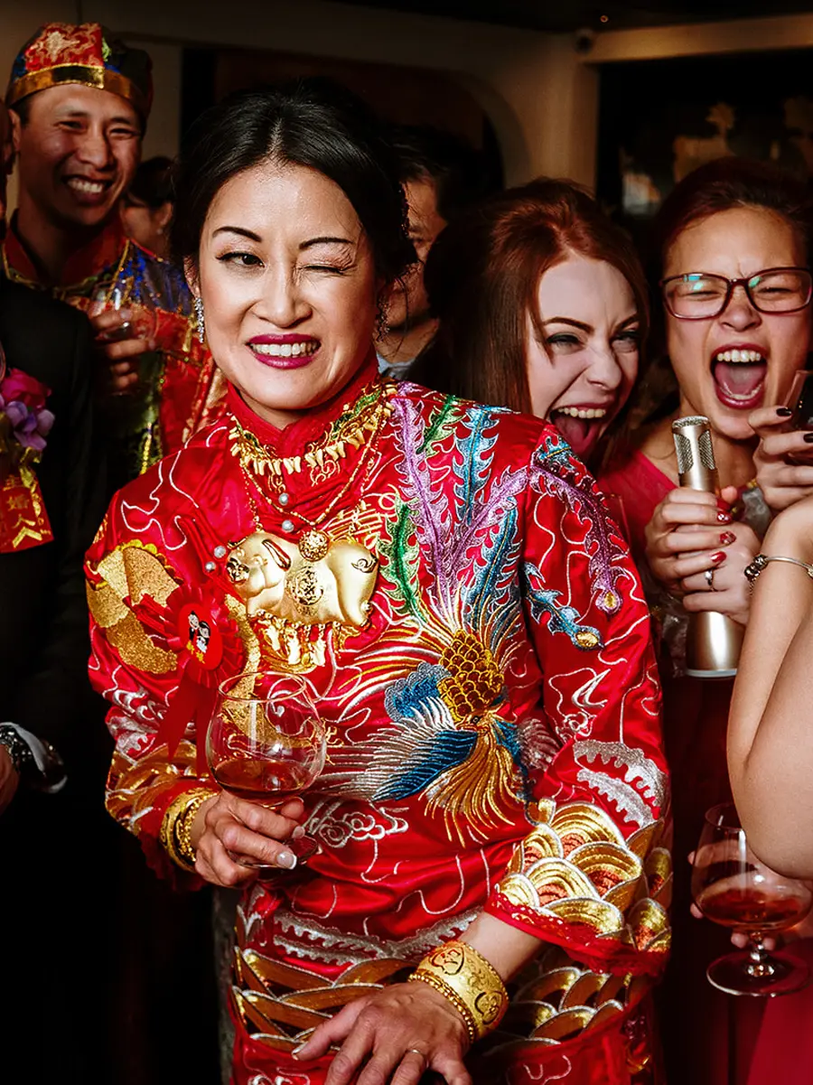Guest in traditional Chinese wedding dress winking at the camera surrounded by laughing guests — documentary wedding photography by Gary Phillip