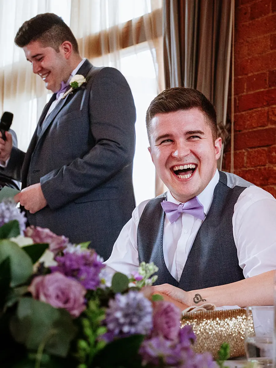 Groom laughing helplessly at the top table during the wedding speeches, best man standing behind him with a microphone — documentary wedding photography by Gary Phillip