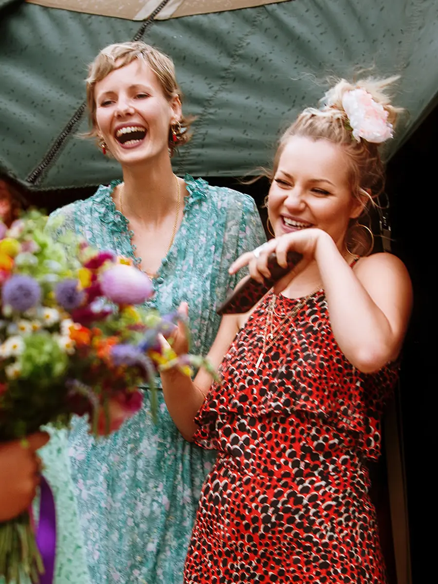 Two female wedding guests laughing together outside a yurt at a festival wedding, wildflower bouquet in hand — documentary wedding photography by Gary Phillip