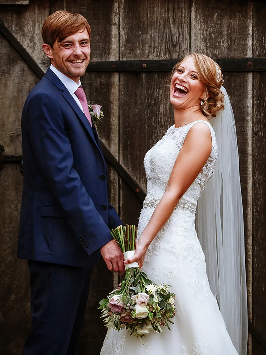 Bride and groom laughing together in front of old barn doors, bride in lace dress holding a blush bouquet — documentary wedding photography by Gary Phillip