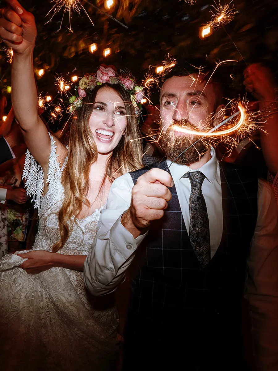 Bride and groom surrounded by sparklers at night, bride in flower crown raising her arm in celebration — documentary wedding photography by Gary Phillip
