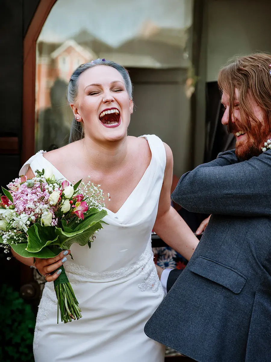 Bride with blue-grey hair laughing with her mouth wide open holding a colourful bouquet, groom smiling beside her — documentary wedding photography by Gary Phillip