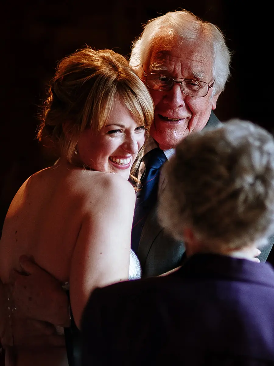 Bride smiling warmly as an elderly gentleman leans in close to her cheek in a dark warmly lit room — documentary wedding photography by Gary Phillip