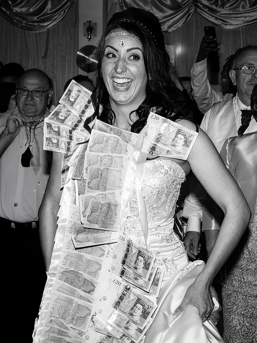 Bride laughing with delight as guests pin twenty pound notes to her wedding dress during a traditional money dance — documentary wedding photography by Gary Phillip