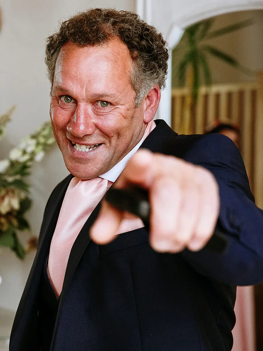 Wedding guest in a navy suit pointing directly at the camera with a wide grin — documentary wedding photography by Gary Phillip