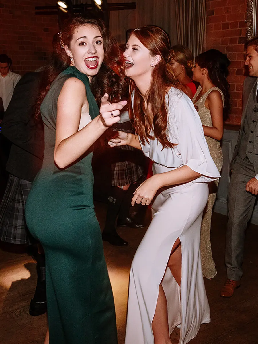 Two female wedding guests dancing together on the dancefloor laughing and pointing at each other in a brick walled venue — documentary wedding photography by Gary Phillip
