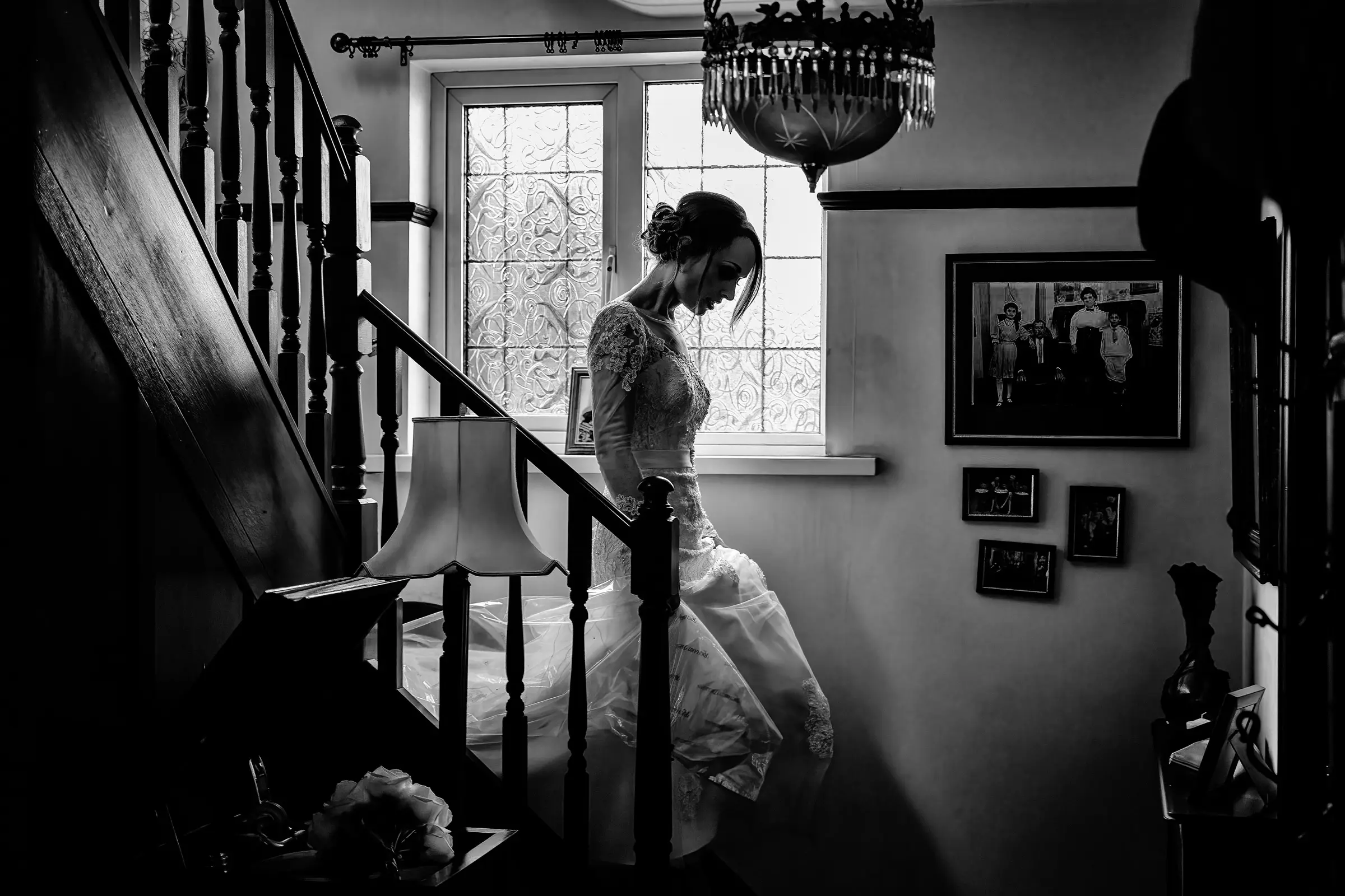 Bride descending a dark wooden staircase in black and white, backlit by a frosted window, wedding dress trailing behind her — documentary wedding photography by Gary Phillip