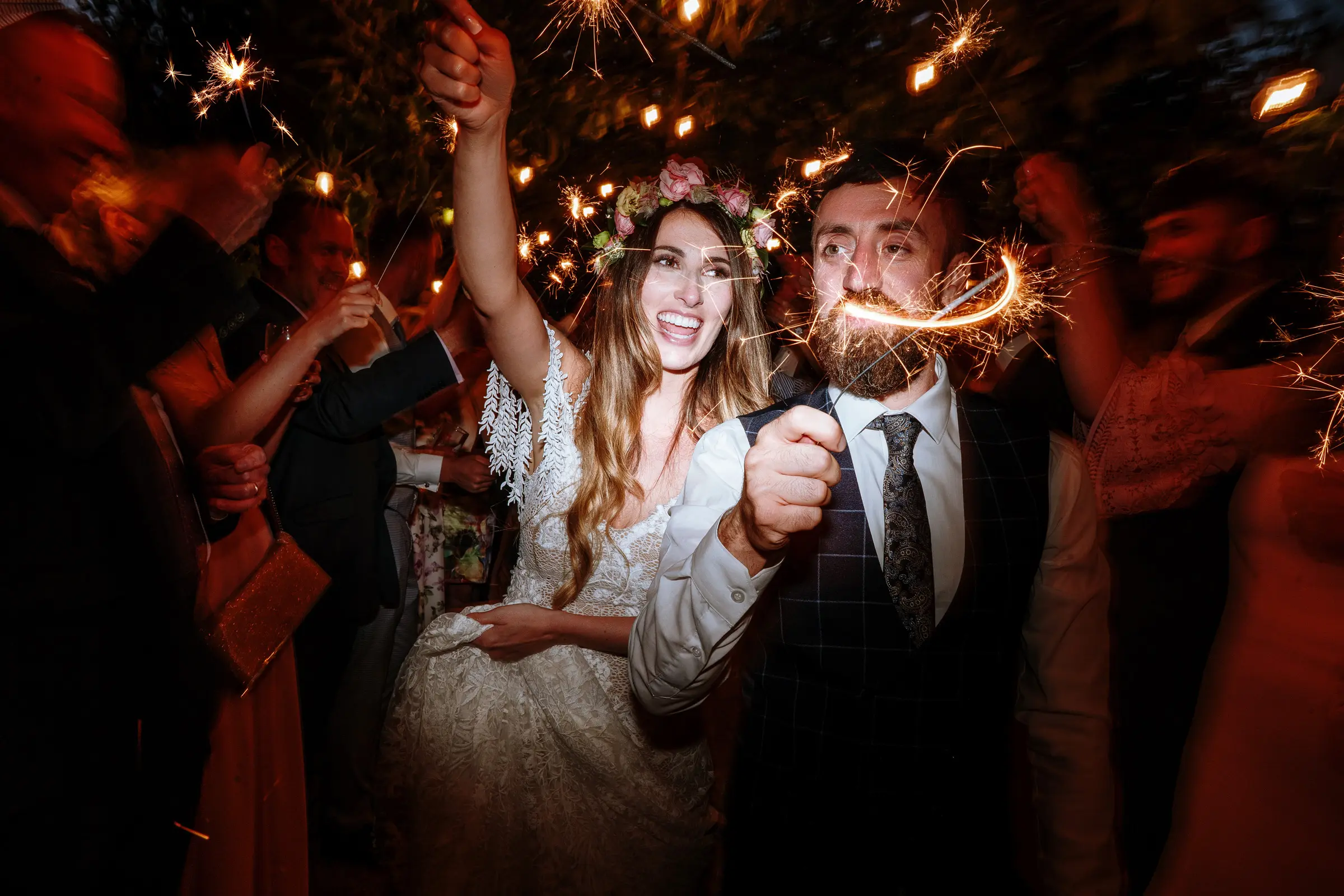 Bride and groom laughing together during a sparkler send-off at night, bride wearing a flower crown, guests and fairy lights surrounding them — documentary wedding photography by Gary Phillip