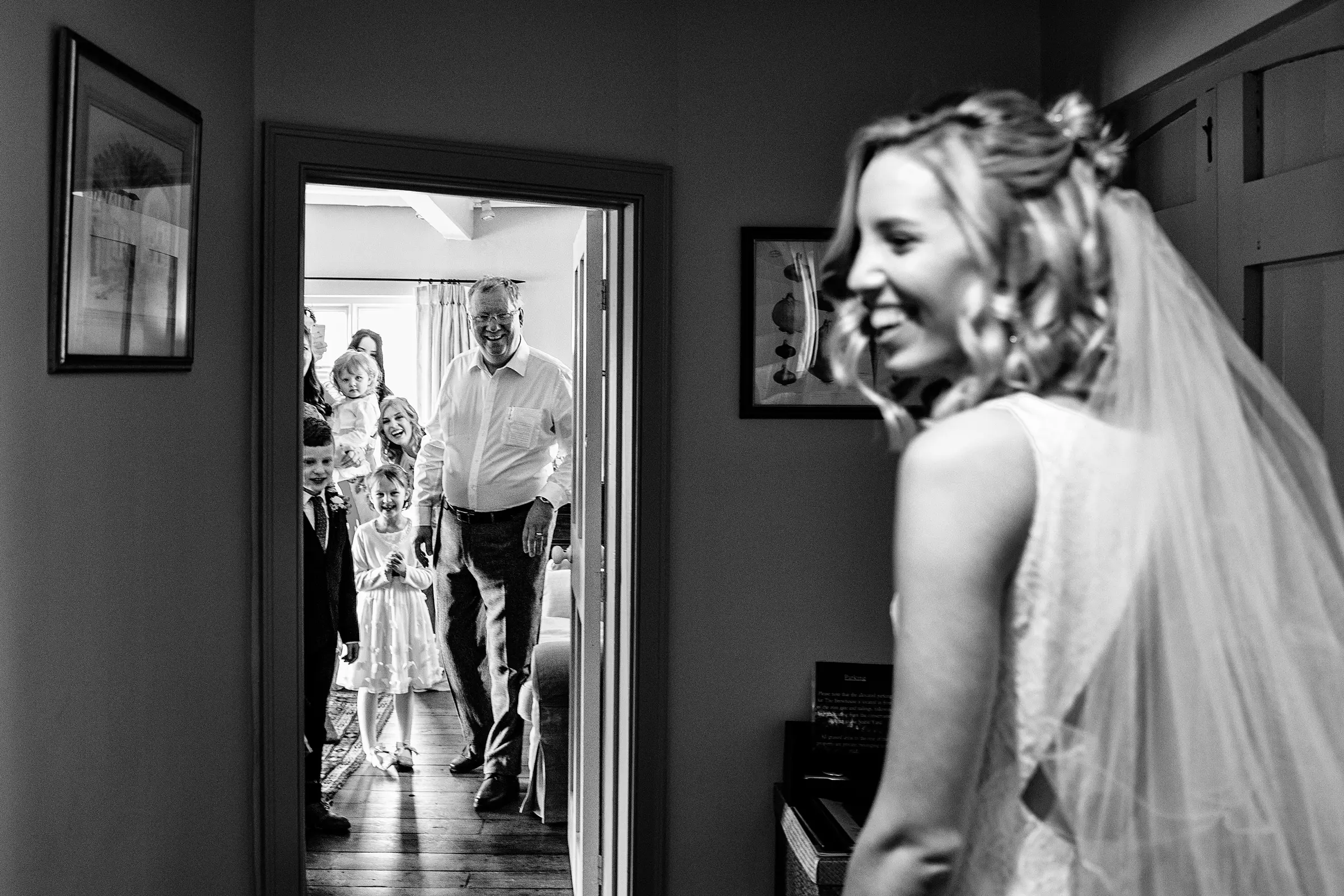 Family and children crowded in bridal suite opening the door and seeing the bride for the first time, father beaming with pride, flower girl clapping with delight — documentary wedding photography by Gary Phillip