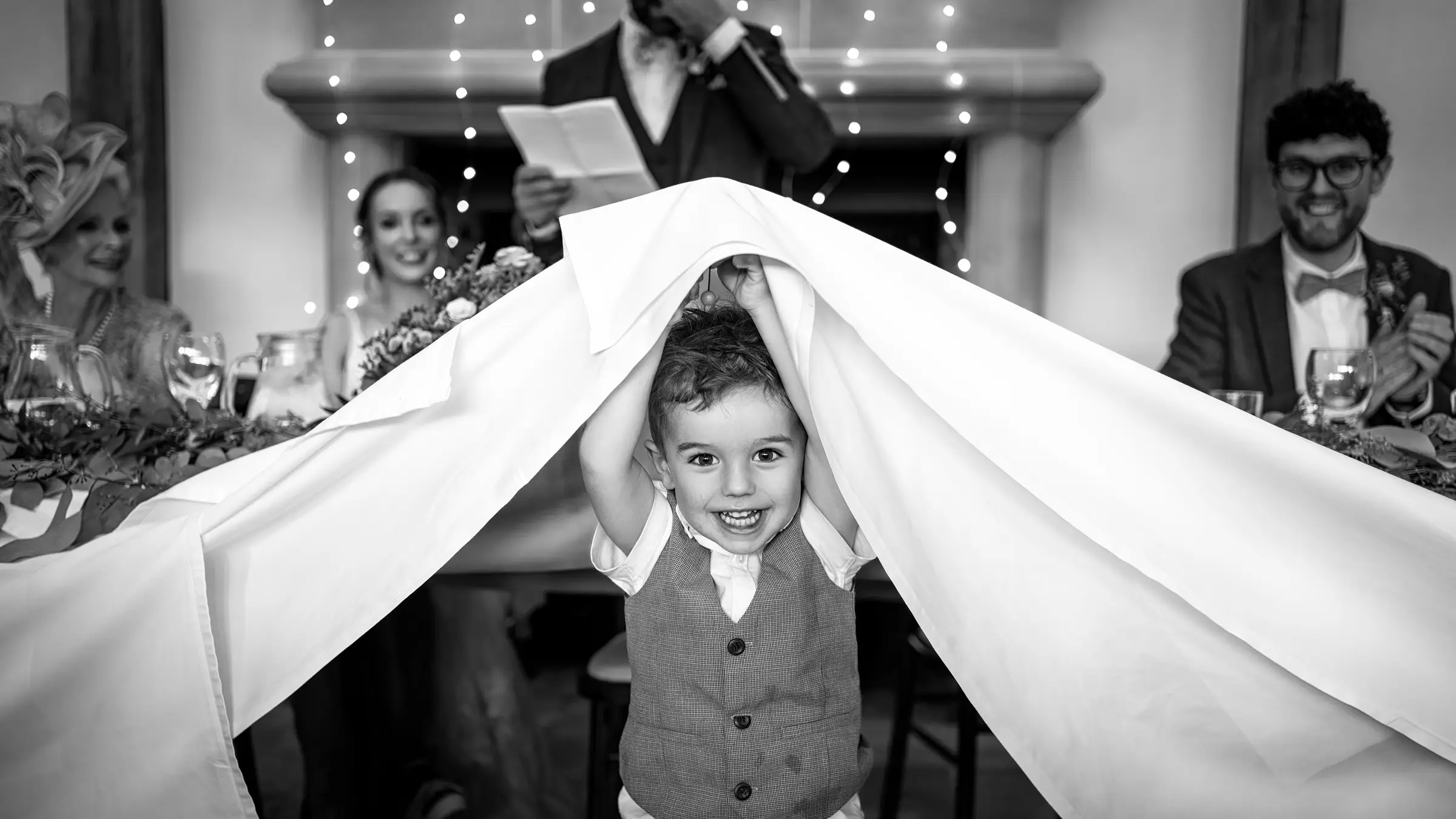 Young boy gleefully lifting the tablecloth over his head during the wedding speeches while the bride laughs and guests look on — documentary wedding photography by Gary Phillip