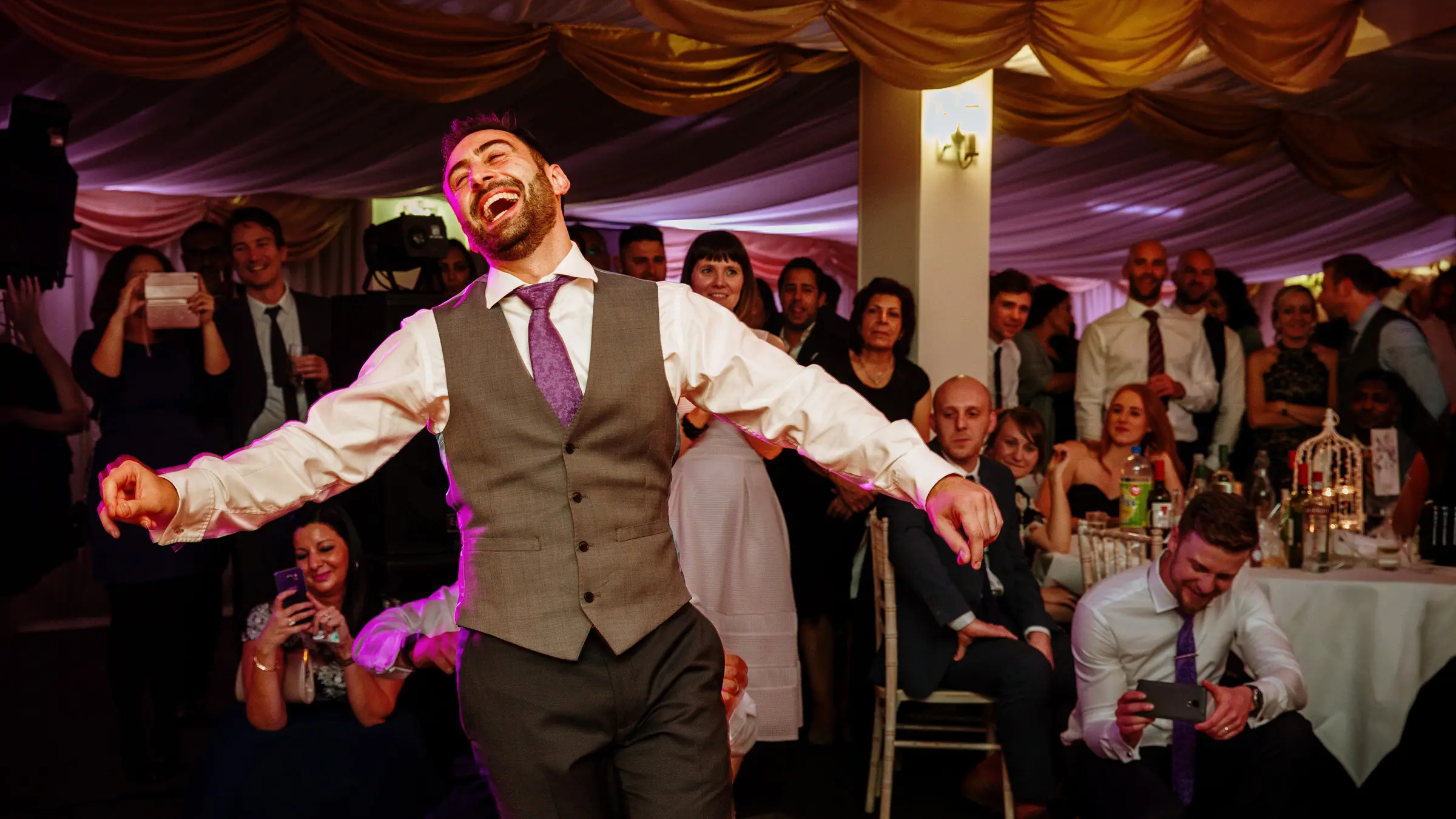 Groom laughing with arms outstretched on the dancefloor, wedding guests watching and cheering from their seats behind him — documentary wedding photography by Gary Phillip