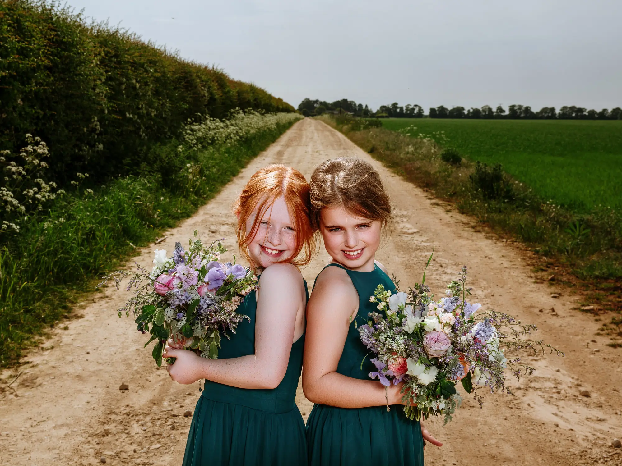 Two young bridesmaids in green dresses holding wildflower bouquets on a long straight country lane, smiling back at the camera — documentary wedding photography by Gary Phillip