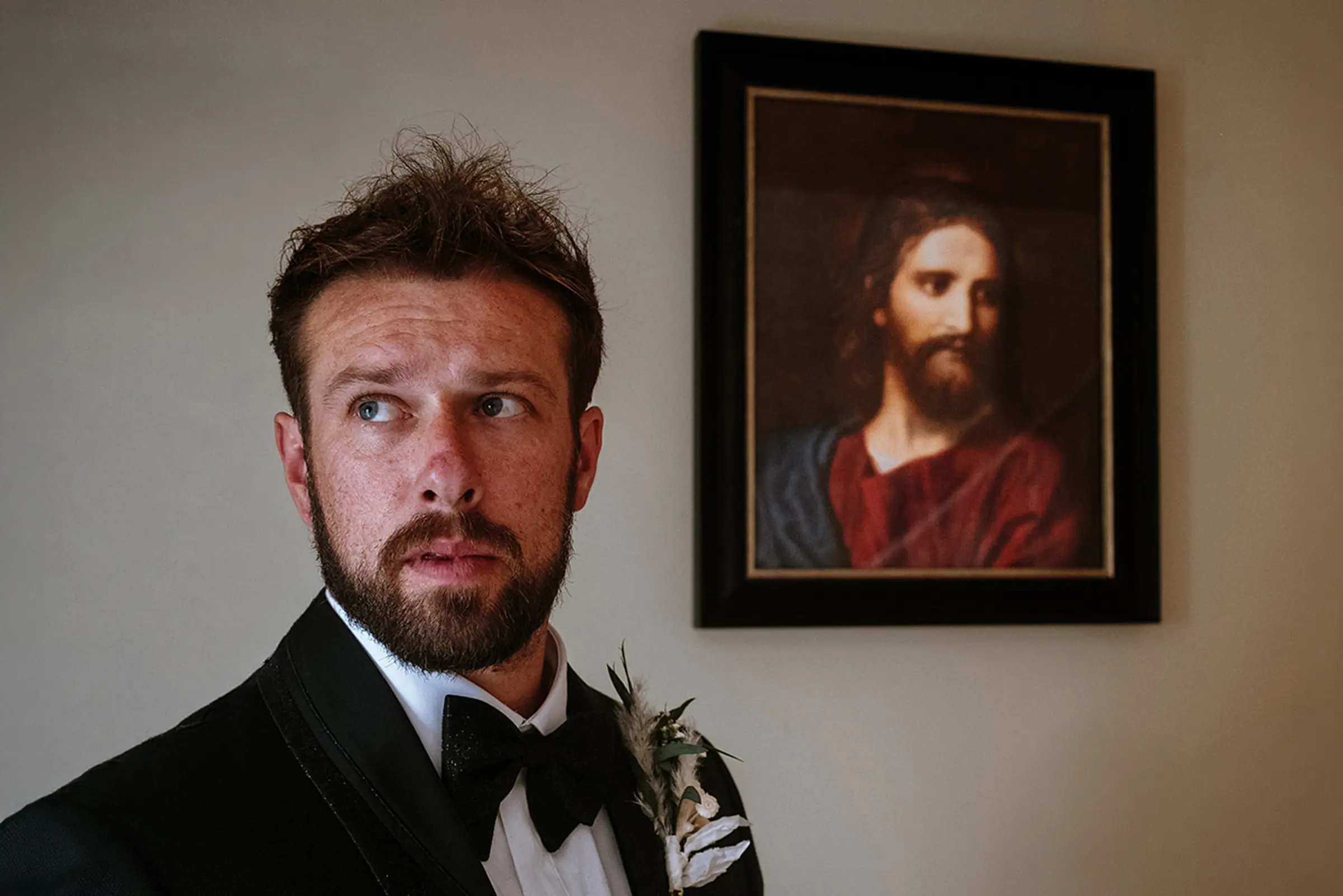 Groom in black tie standing in front of a portrait of Jesus on the wall, looking sideways with a calm intensity — documentary wedding photography by Gary Phillip