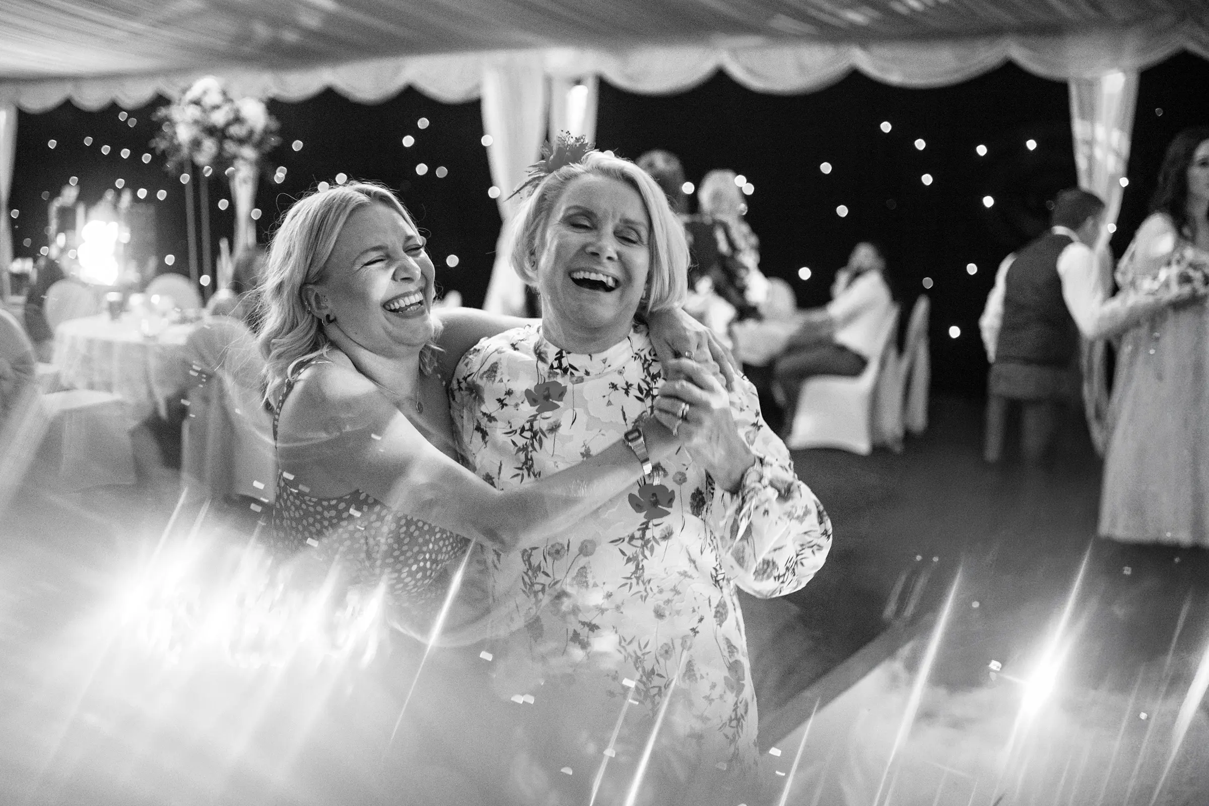 Two women laughing and dancing together on the wedding dancefloor at night, fairy lights and lens flare surrounding them — documentary wedding photography by Gary Phillip
