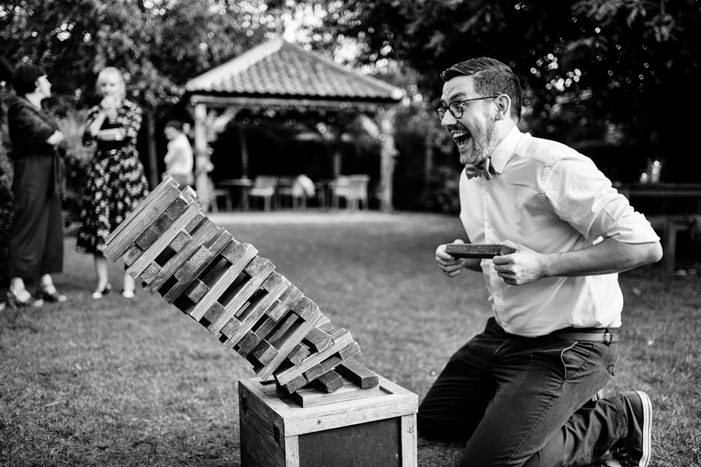 Wedding guest in glasses reacting with delight as a giant Jenga tower topples over in the garden during the drinks reception — documentary wedding photography by Gary Phillip