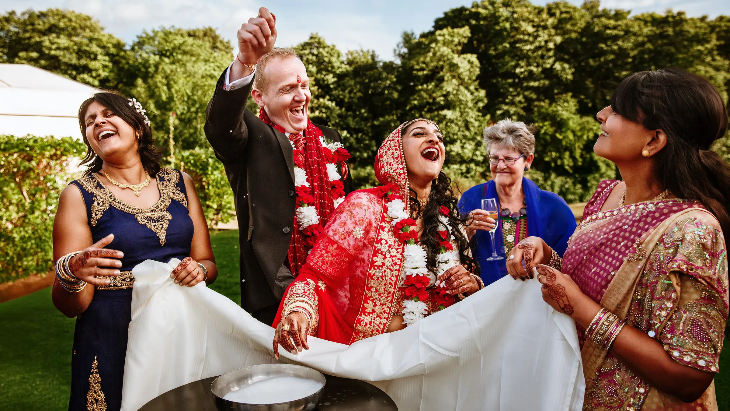 Hindu-Western wedding ceremony outdoors, bride in red and gold lehenga laughing joyfully with groom and family during a traditional ritual — documentary wedding photography by Gary Phillip