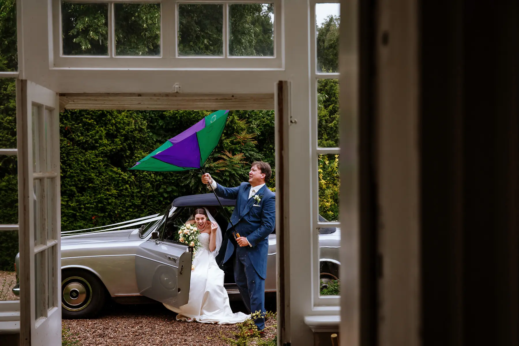 Bride stepping out of a vintage wedding car in the rain while a groomsman wrestles with an inside-out umbrella, both laughing — documentary wedding photography by Gary Phillip