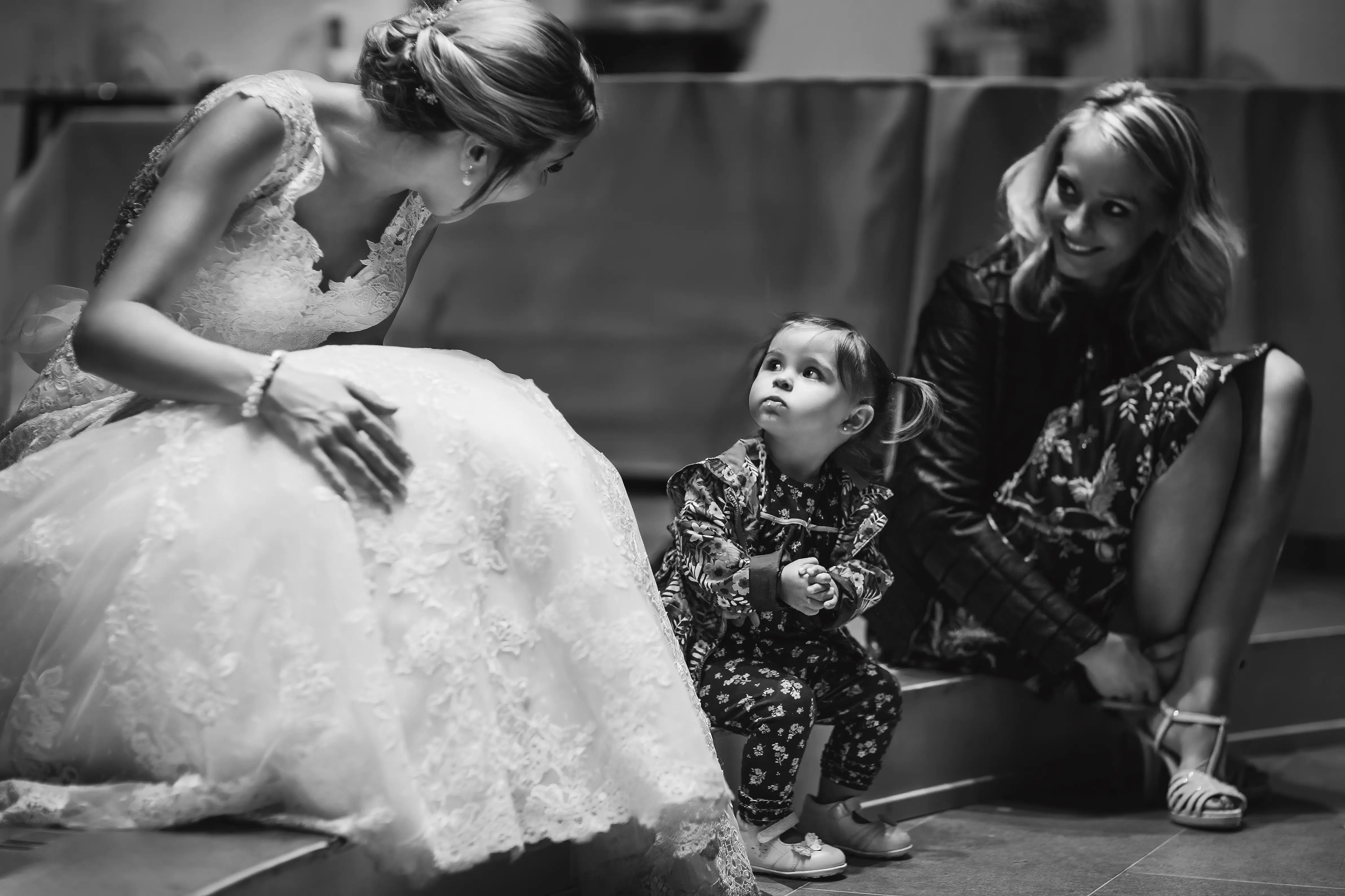 Bride in full lace wedding dress crouching down to speak to a small child at eye level, a tender quiet moment — documentary wedding photography by Gary Phillip