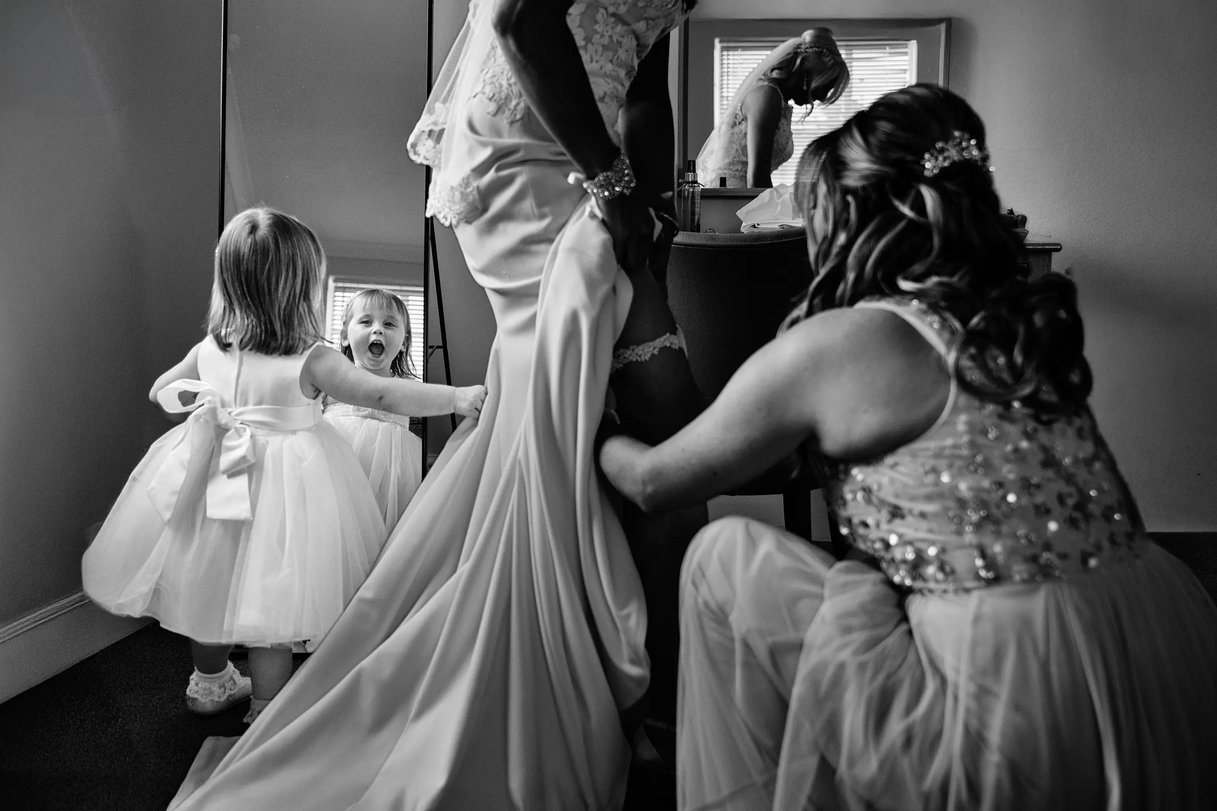Flower girl staring open-mouthed with wonder at herself while a bridesmaid fits the bride's garter during wedding morning preparations, captured in black and white — documentary wedding photography by Gary Phillip