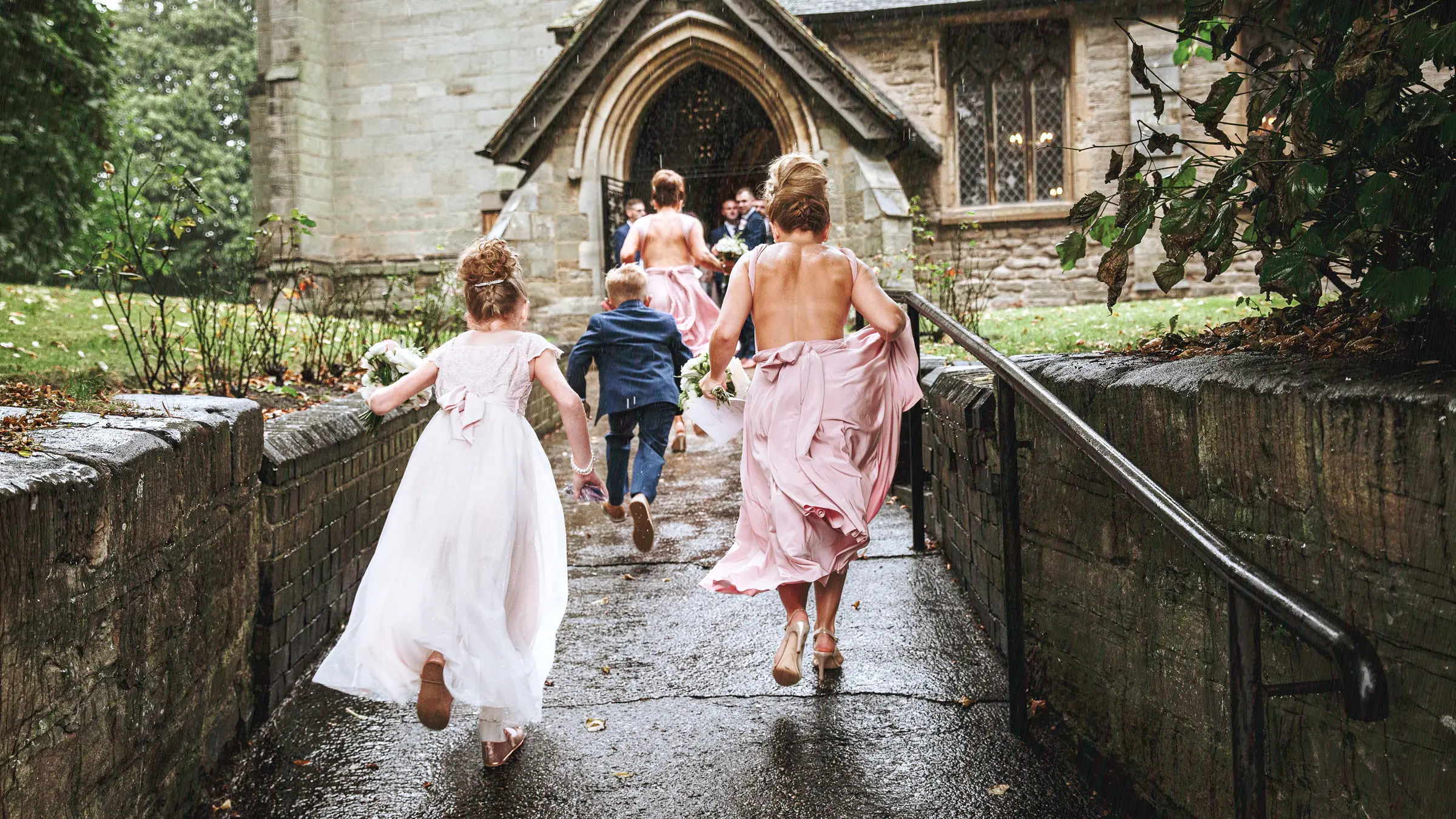 Bridesmaids and flower girl running through the rain toward a Gothic stone church entrance, dresses flying — documentary wedding photography by Gary Phillip