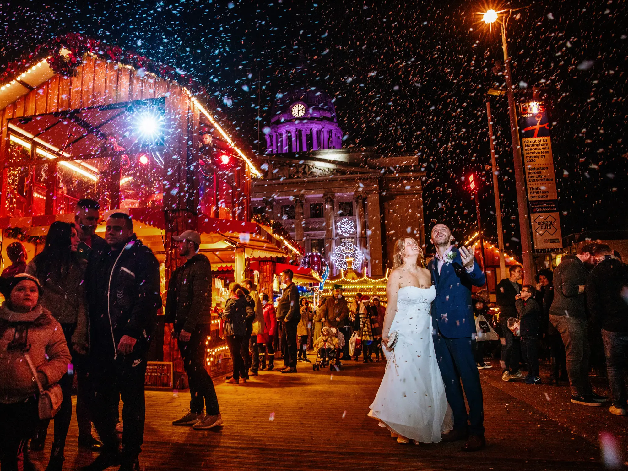 Bride and groom standing in falling snow at Nottingham Christmas market at night, Council House lit up behind them — documentary wedding photography by Gary Phillip