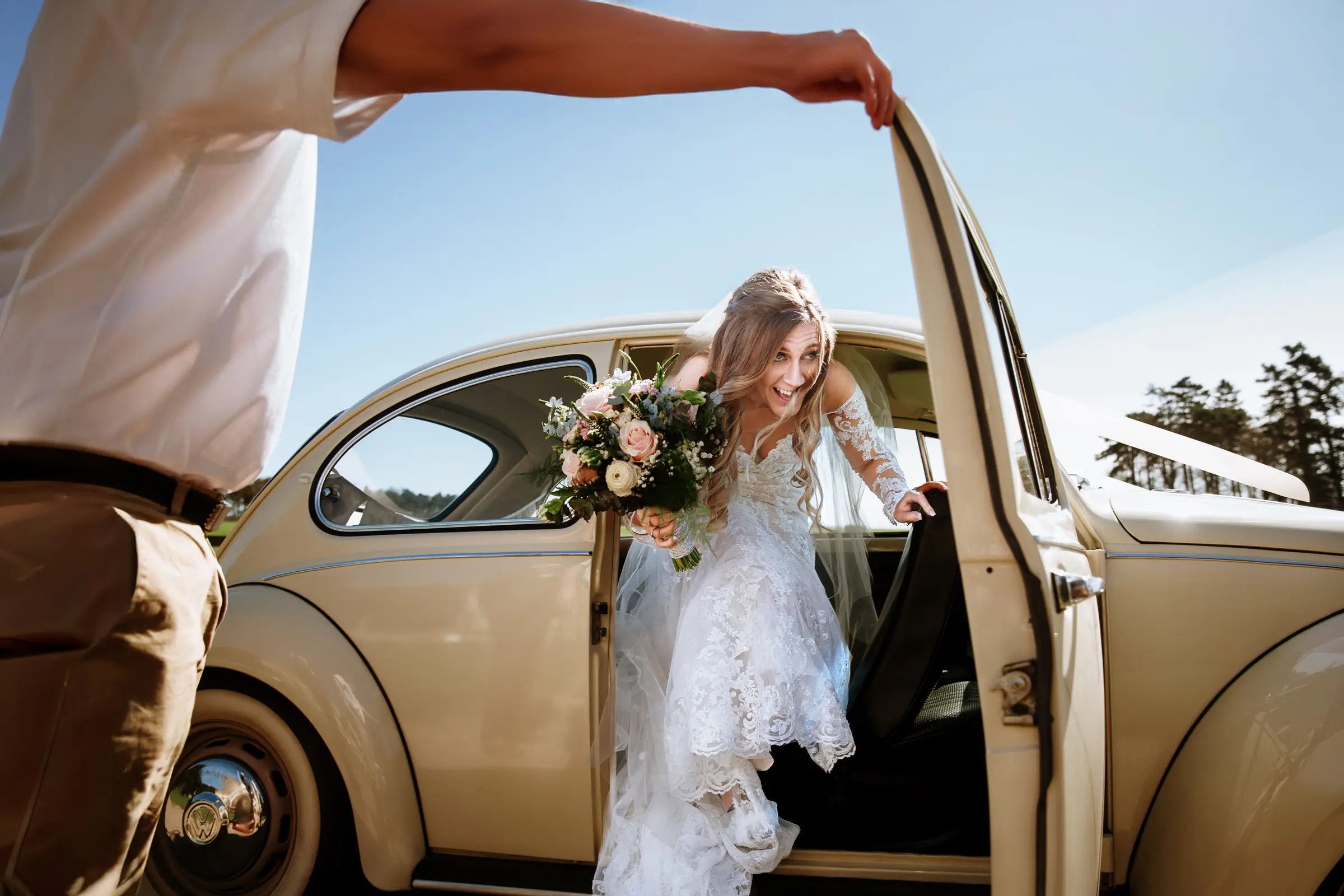 Bride stepping out of a cream vintage VW Beetle on a sunny wedding day, laughing as the door is held open — documentary wedding photography by Gary Phillip