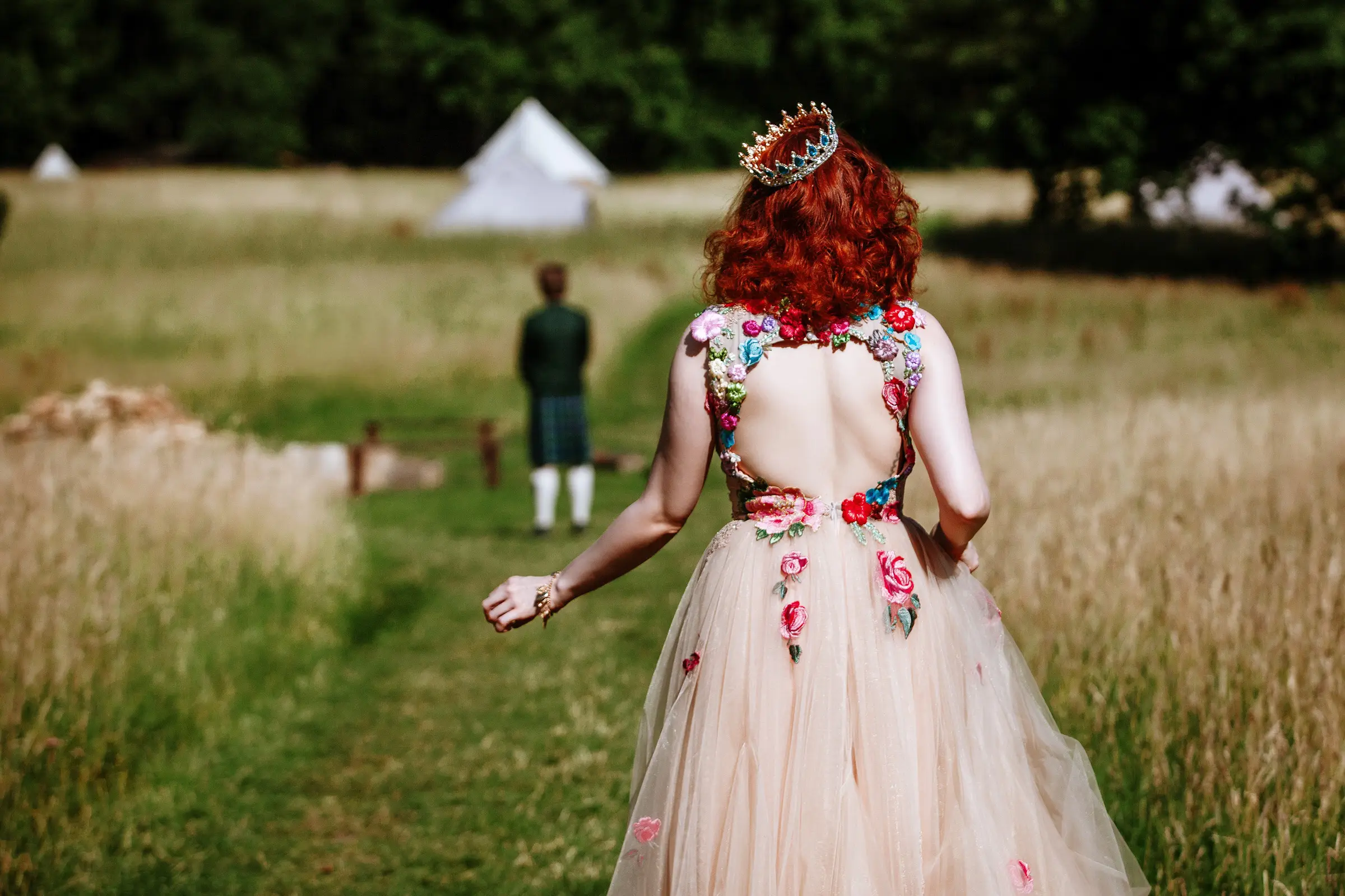 Bride going to her first look, she has red hair and a colourful floral embroidered open-back dress walking toward her groom across a sunlit field, glamping tents in the distance — documentary wedding photography by Gary Phillip