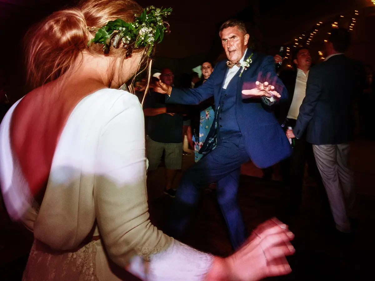 Father of the bride dancing energetically on the dancefloor facing the bride, blue suit and floral bow tie, fairy lights behind them — documentary wedding photography by Gary Phillip