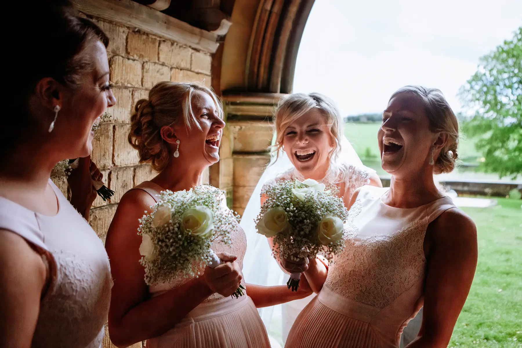 Bride and bridesmaids laughing together outside a stone church doorway, all holding white rose and gypsophila bouquets — documentary wedding photography by Gary Phillip