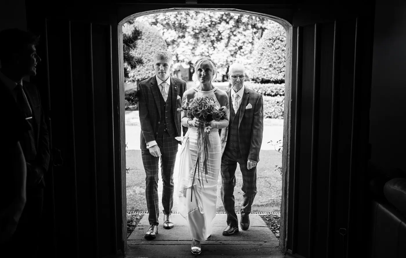 Bride Amy moving through the coach house doorway at Doddington Hall with her son and father following behind on her wedding day