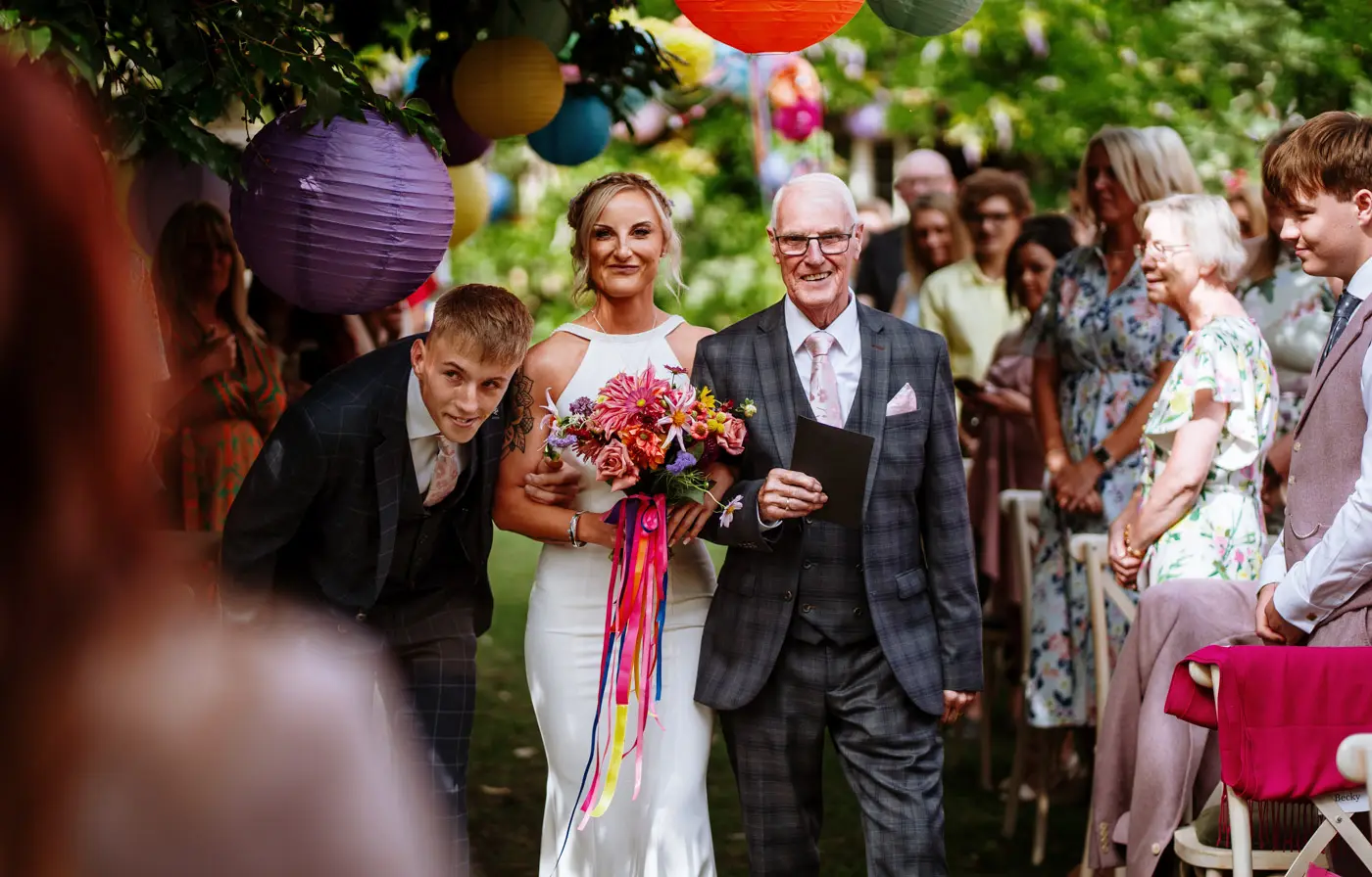 Amy and her family approaching the front of the ceremony room at Doddington Hall, all smiling as they see Abby while Amy's son ducks under the colourful hanging lanterns