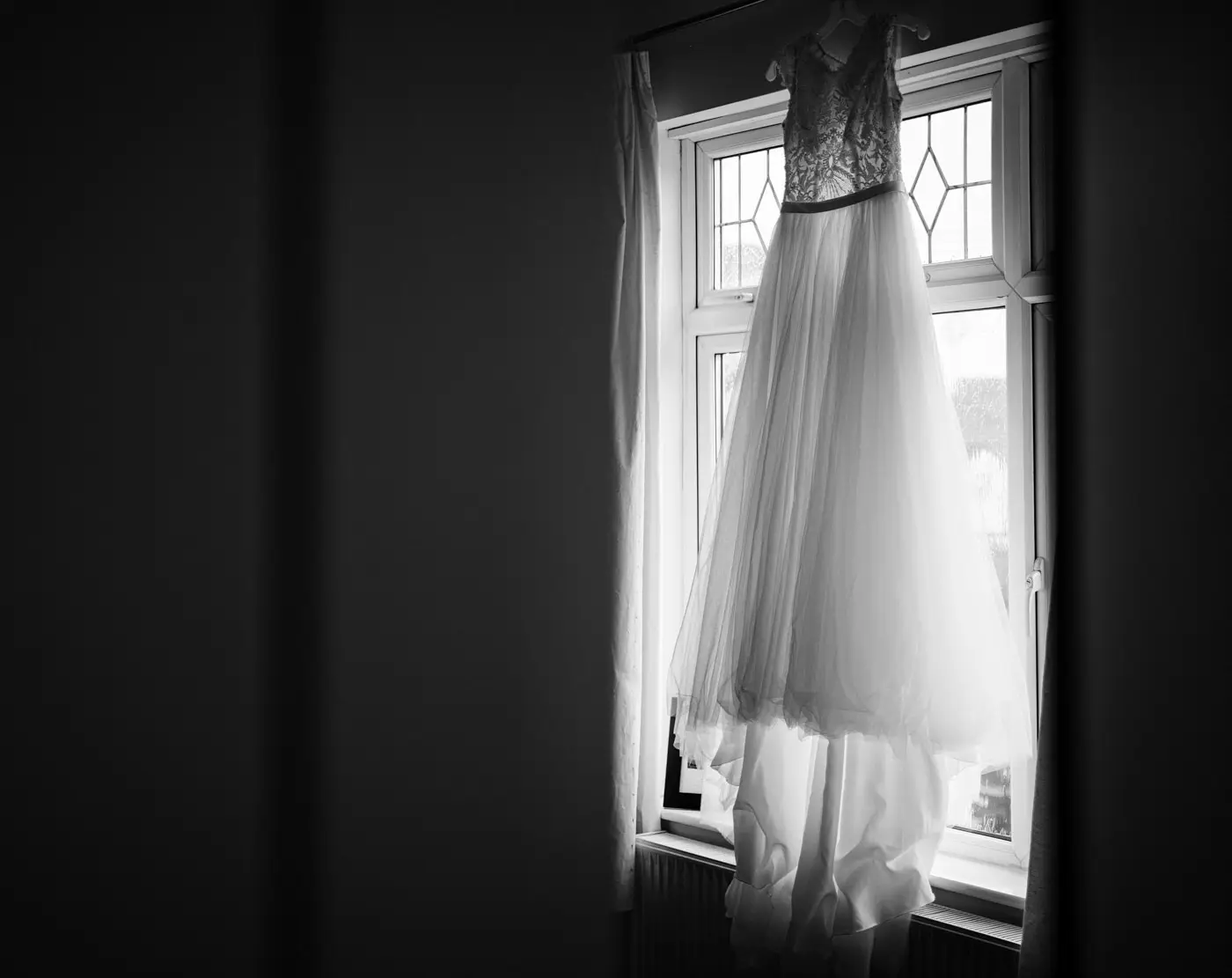 An elegant A-line wedding gown hanging in a window on the morning of Ellen and Ben's wedding at Carriage Hall, Nottinghamshire