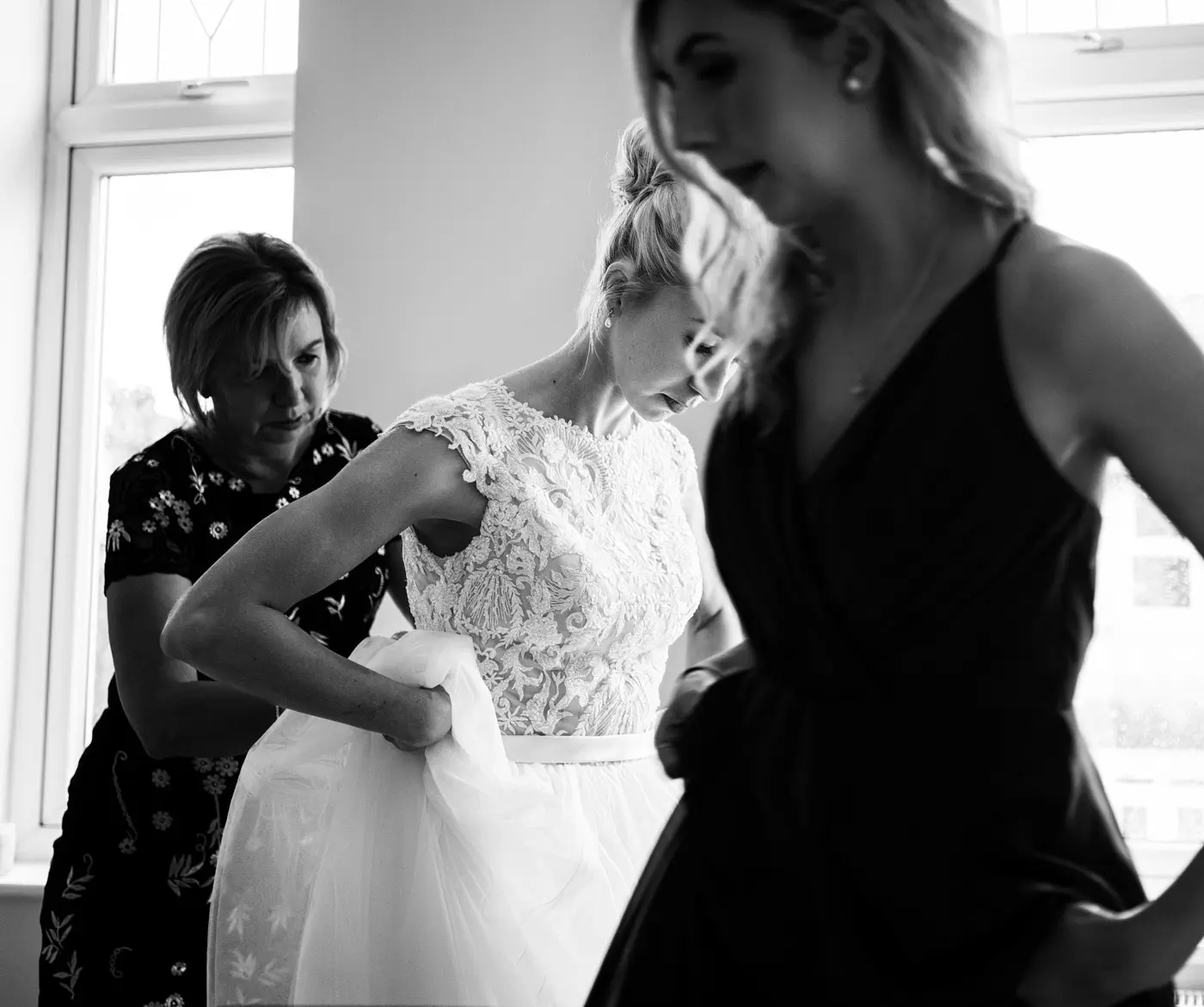 Black and white square crop of bride Ellen centred between her mum and chief bridesmaid as she gets dressed on her wedding morning