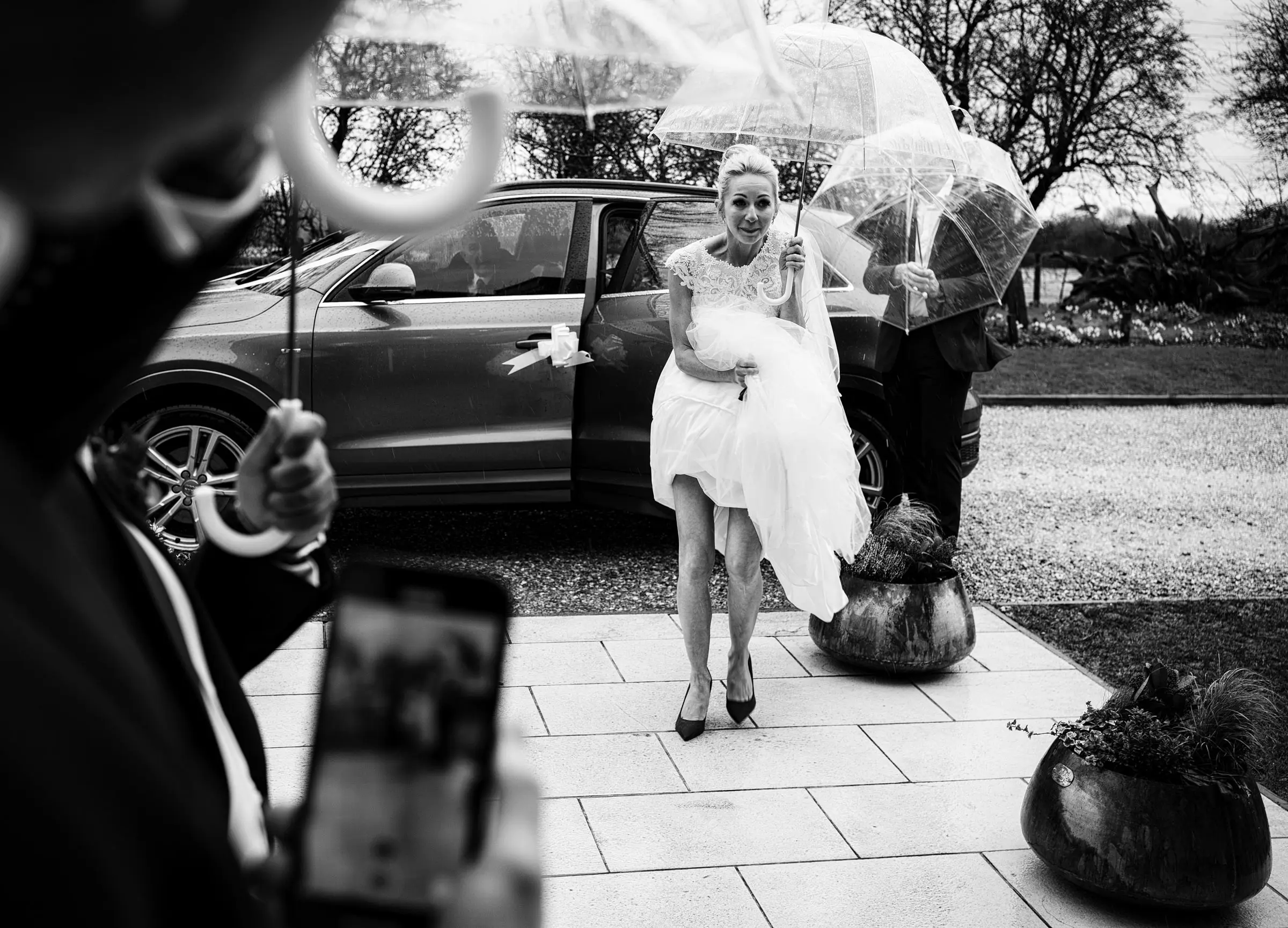 Bride Ellen emerging from the wedding car in the pouring February rain with her dress hiked up to avoid the water at Carriage Hall, Nottinghamshire