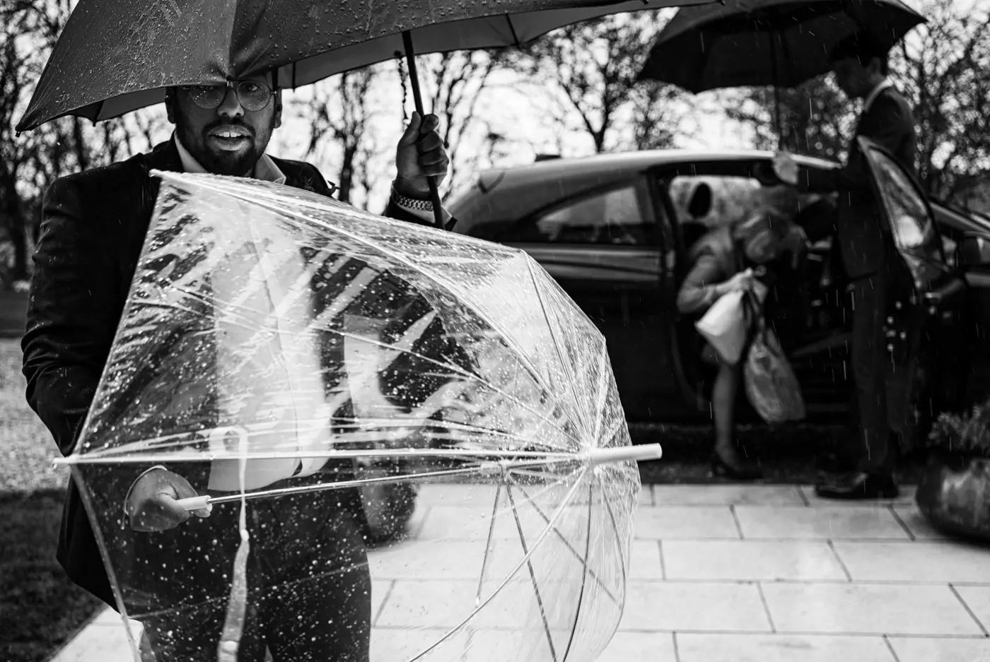 Best man awaiting guests with two umbrellas while another groomsman helps a guest out of a car at Carriage Hall, Nottinghamshire