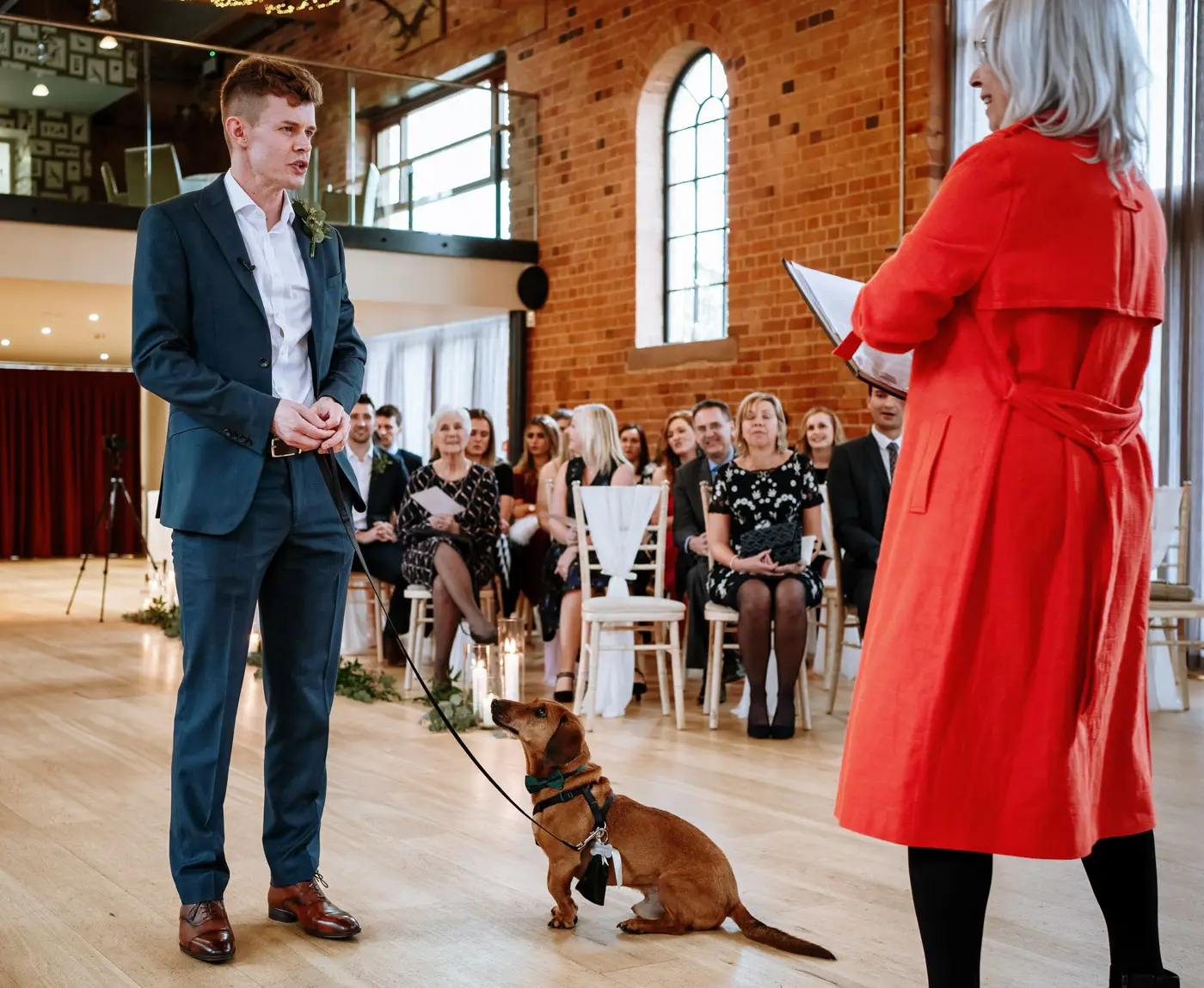 Groom Ben waiting at the front of the ceremony room at Carriage Hall with the sausage dog ring bearer sitting beside him