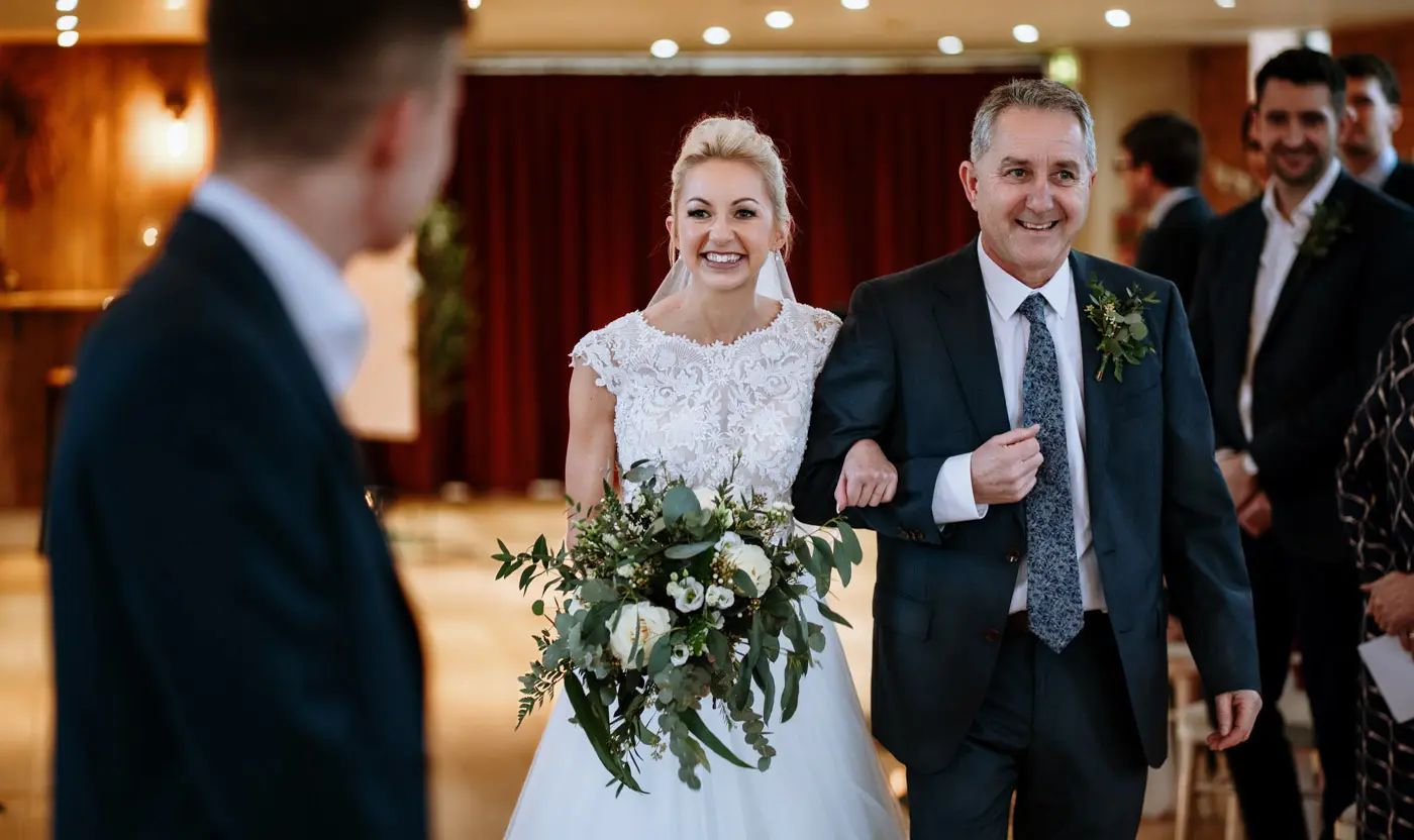 Bride Ellen smiling at groom Ben as she approaches the front of the ceremony room at Carriage Hall, Nottinghamshire