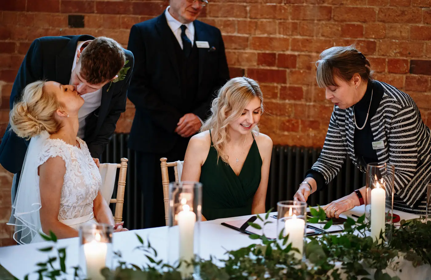 A bridesmaid signs the wedding register at Carriage Hall as Ellen and Ben share a kiss in the background, Nottinghamshire