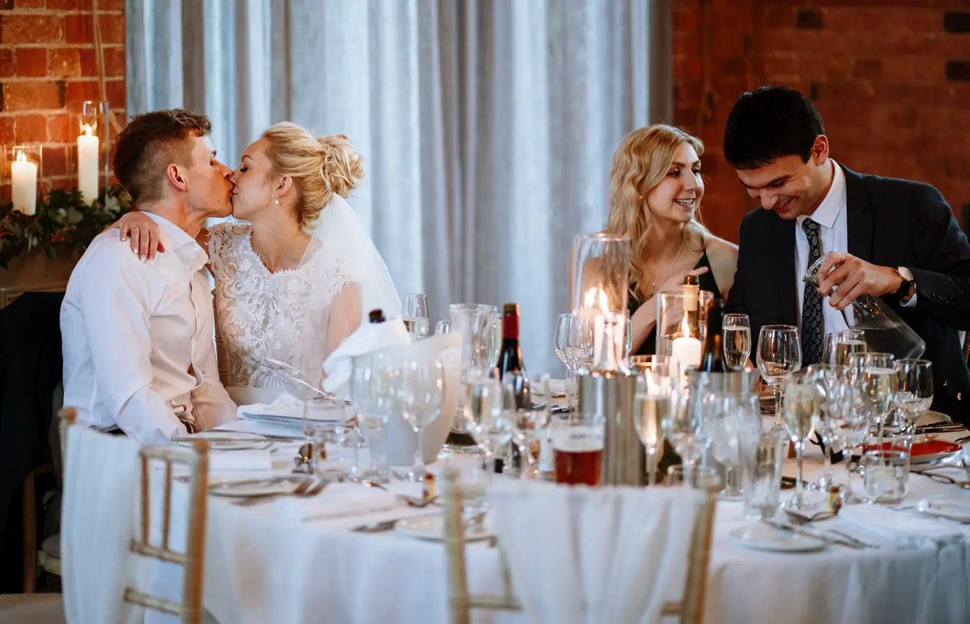 Ellen and Ben sharing a kiss at the top table at Carriage Hall while a bridesmaid and her partner chat beside them