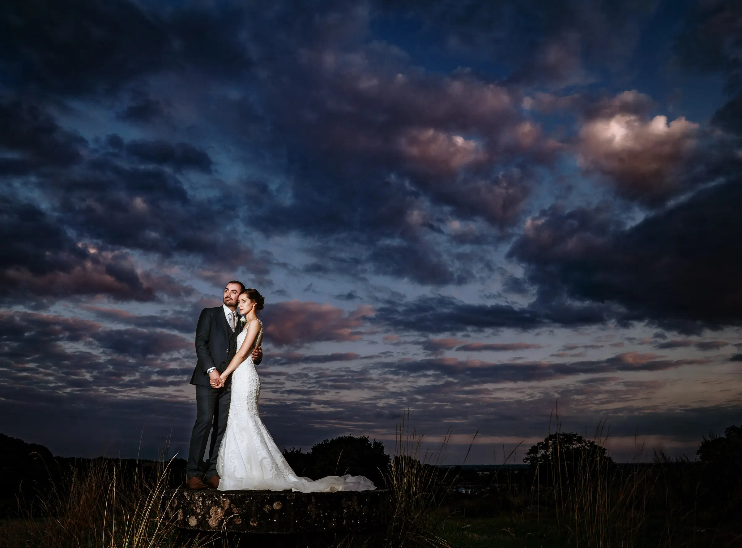 Late evening portrait of bride Krissie and groom Tom lit against a dramatic dark blue sky with pink clouds at Swancar Farm, Nottinghamshire, documentary wedding photography by Gary Phillip