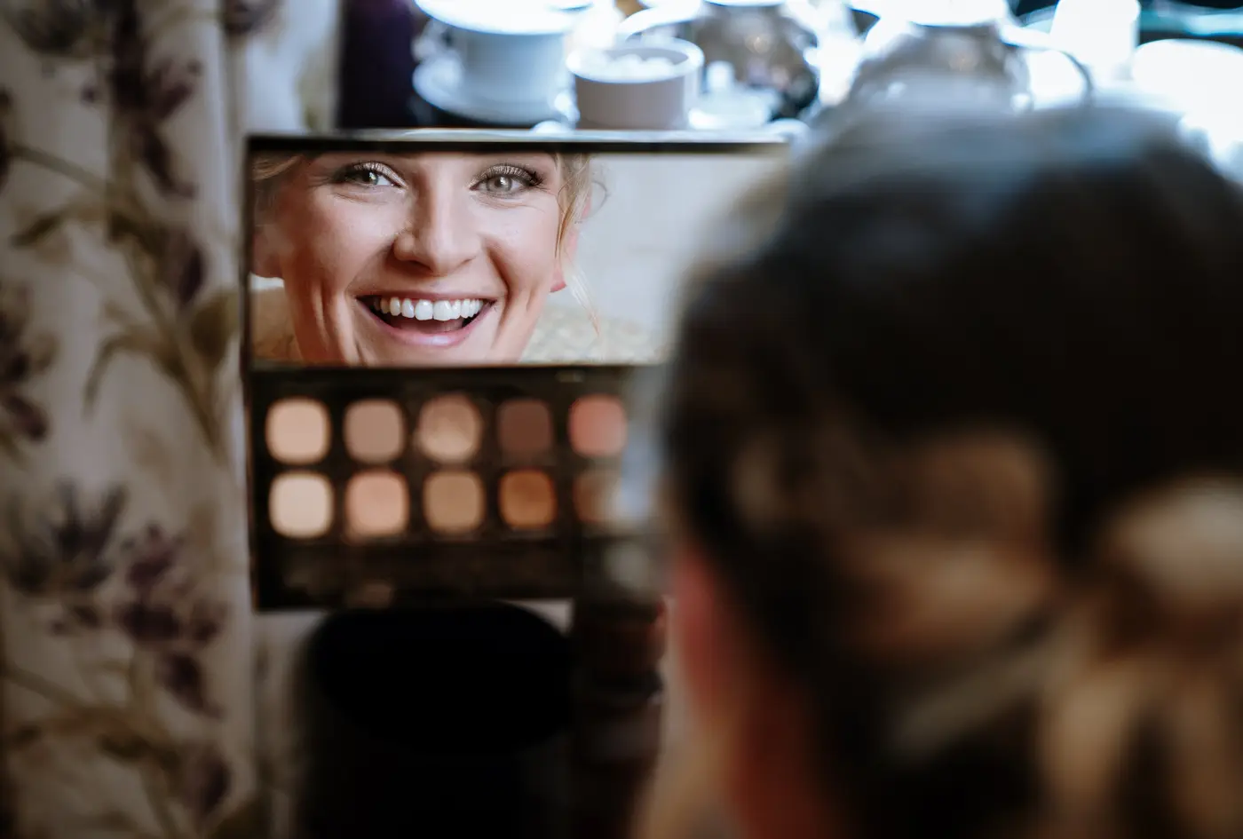 Bridesmaid laughing at her own reflection in a mirror while applying her makeup on the wedding morning at Beeston Fields, Nottingham