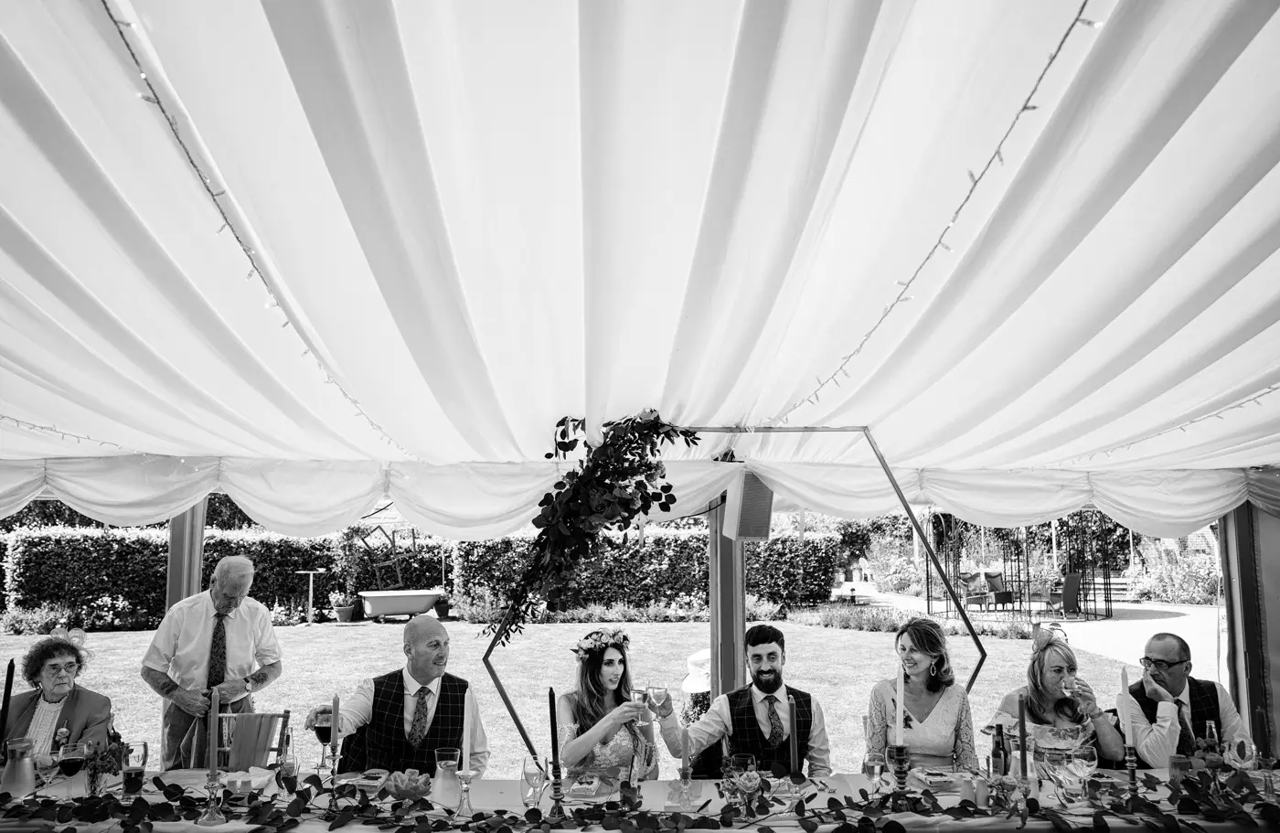 Black and white view of the full top table at Beeston Fields with bride Laura and groom Gavin clinking glasses in a toast while conversations happen all around them