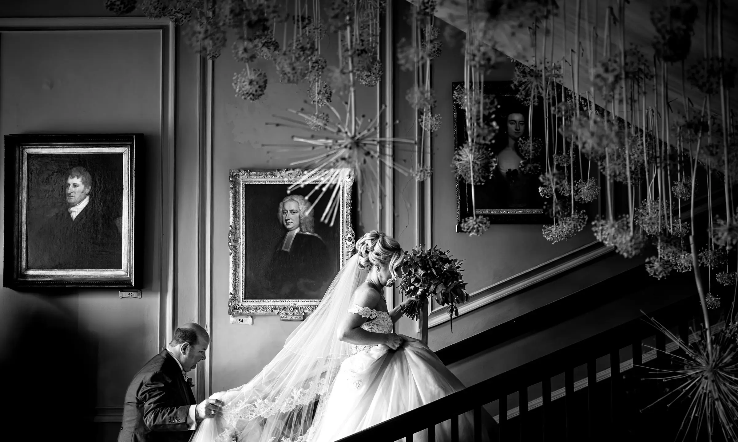 Black and white image of bride Louise ascending the grand staircase at Doddington Hall past the historic oil portraits of past family members, Lincolnshire