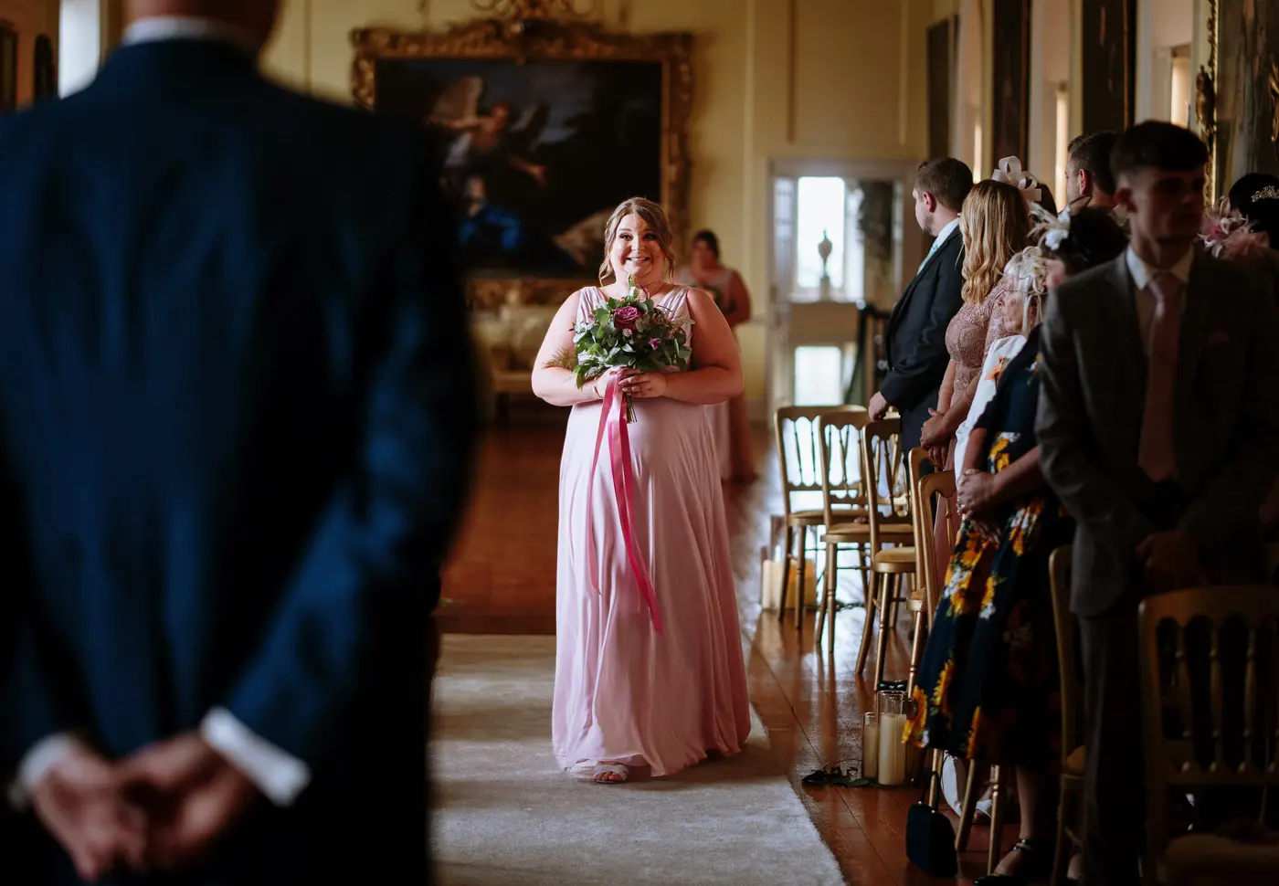 A bridesmaid making her way down the aisle in the Long Gallery at Doddington Hall with a big grin on her face before Louise and Martin's ceremony