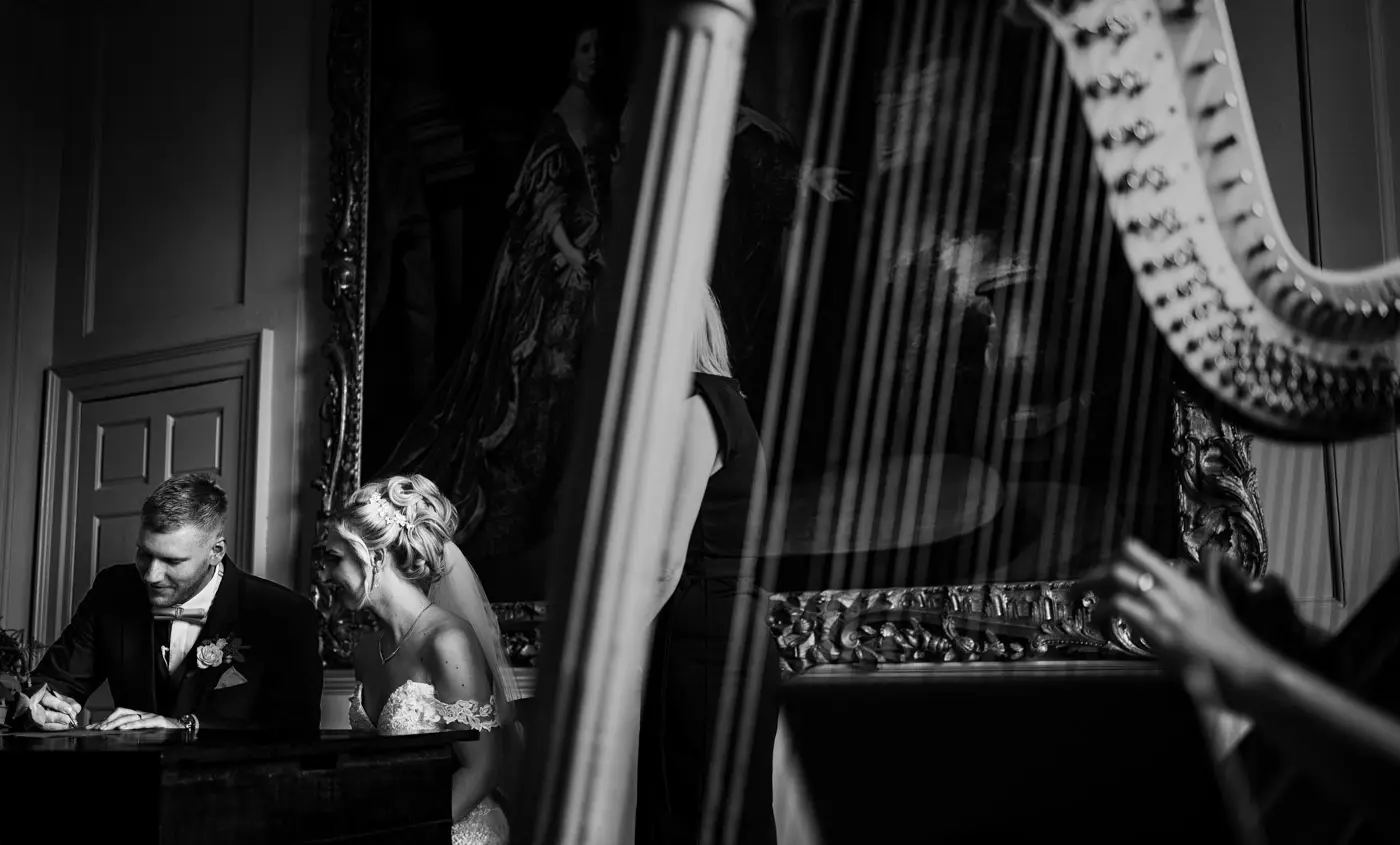 Bride Louise and groom Martin signing the wedding register at Doddington Hall with the harpist visible in the foreground, Lincolnshire