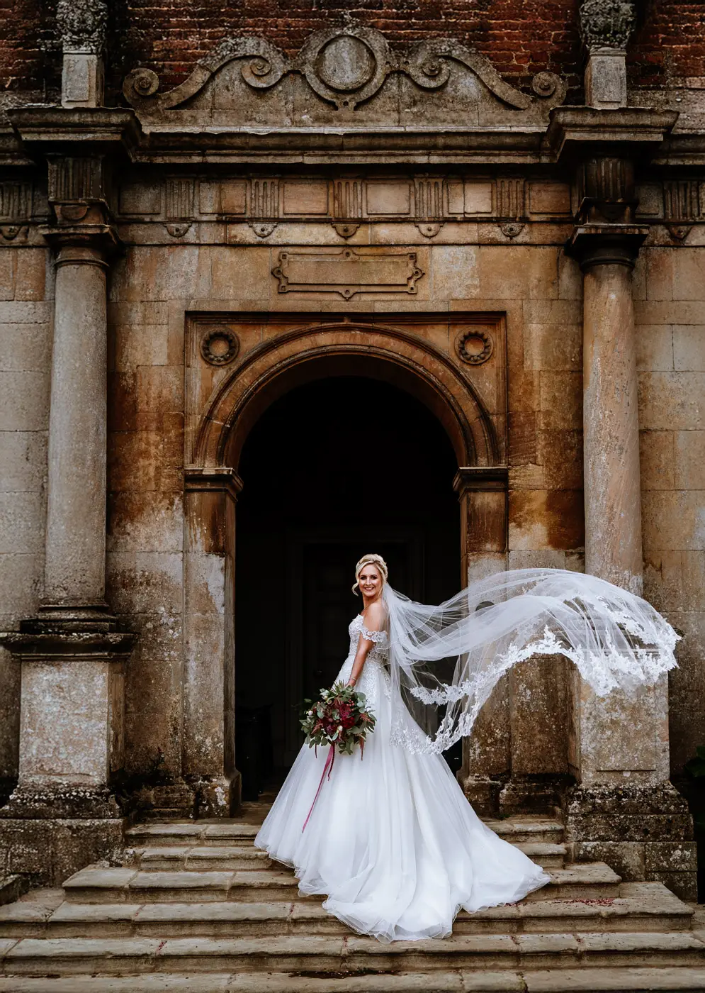 Bride Louise with her cathedral veil flowing in front of the main door at Doddington Hall, a beautiful natural bridal portrait by Gary Phillip