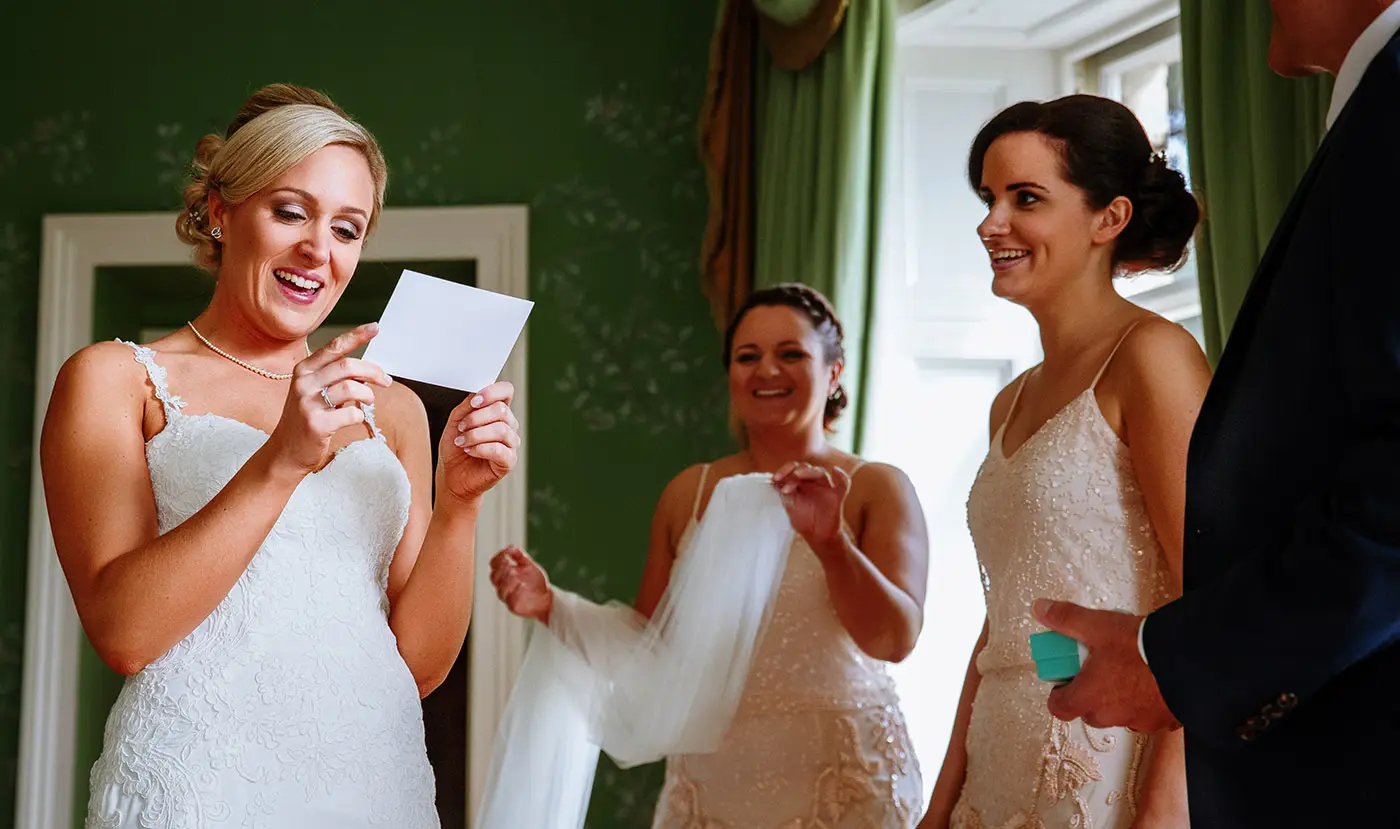 Bride Sally reading a note sent from groom Luke before the ceremony at Stubton Hall while her father and bridesmaids look on with big smiles, documentary wedding photography by Gary Phillip