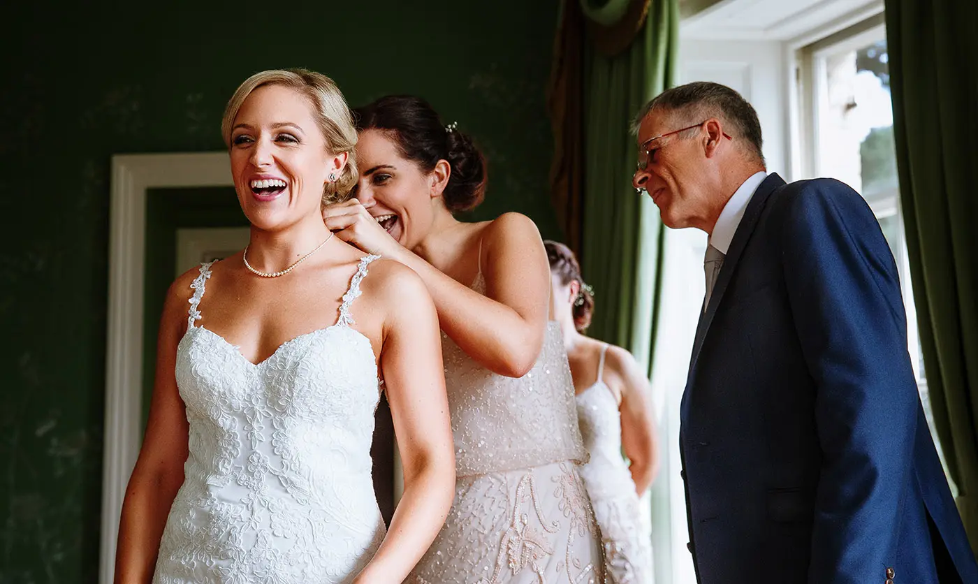 Bridesmaid fastening bride Sally's necklace with big smiles all round at Stubton Hall, Nottinghamshire, a warm wedding morning moment by Gary Phillip