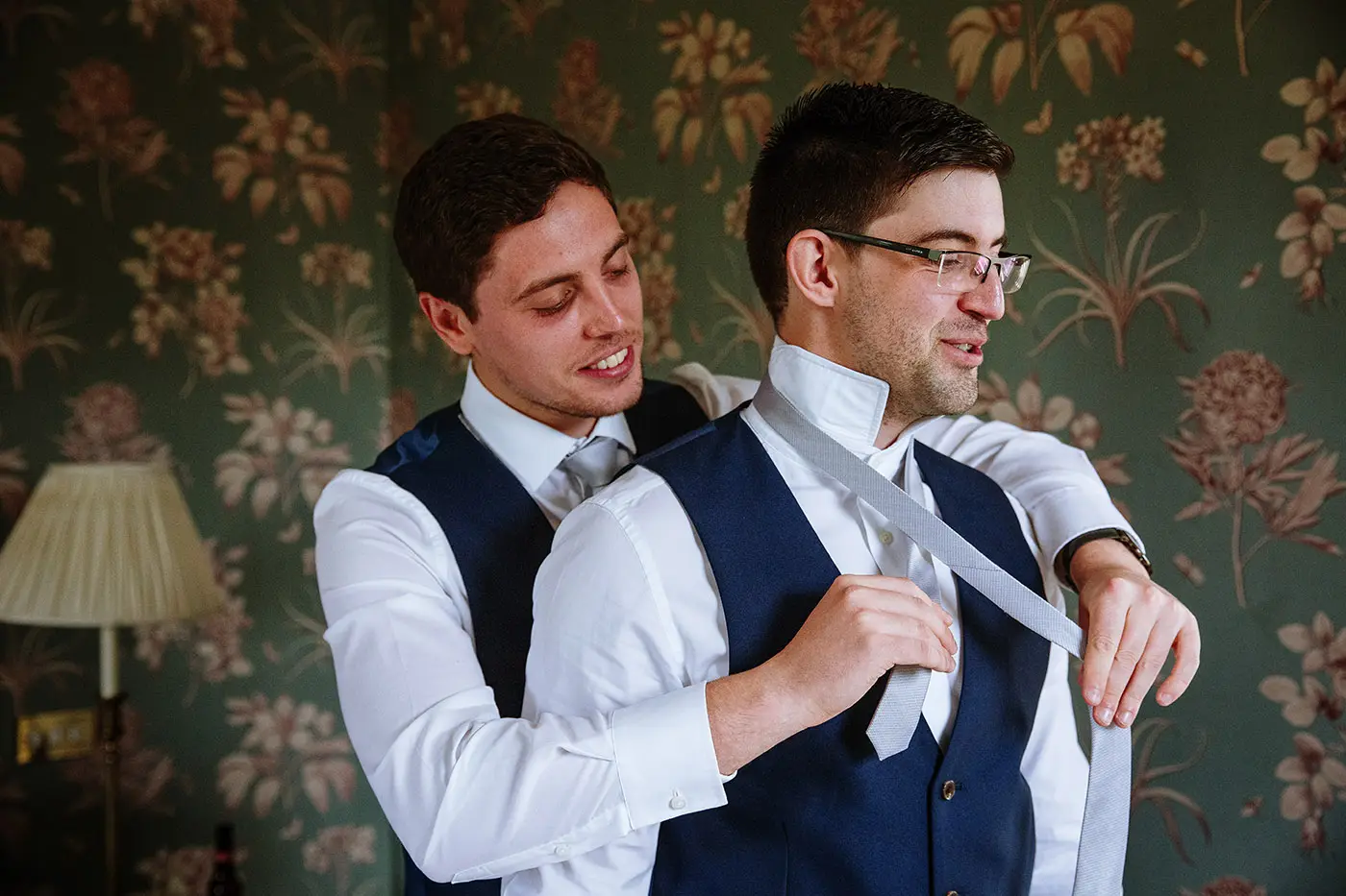 One groomsman helping another tie his tie from behind at Stubton Hall, Nottinghamshire, a relaxed and humorous wedding morning moment by Gary Phillip